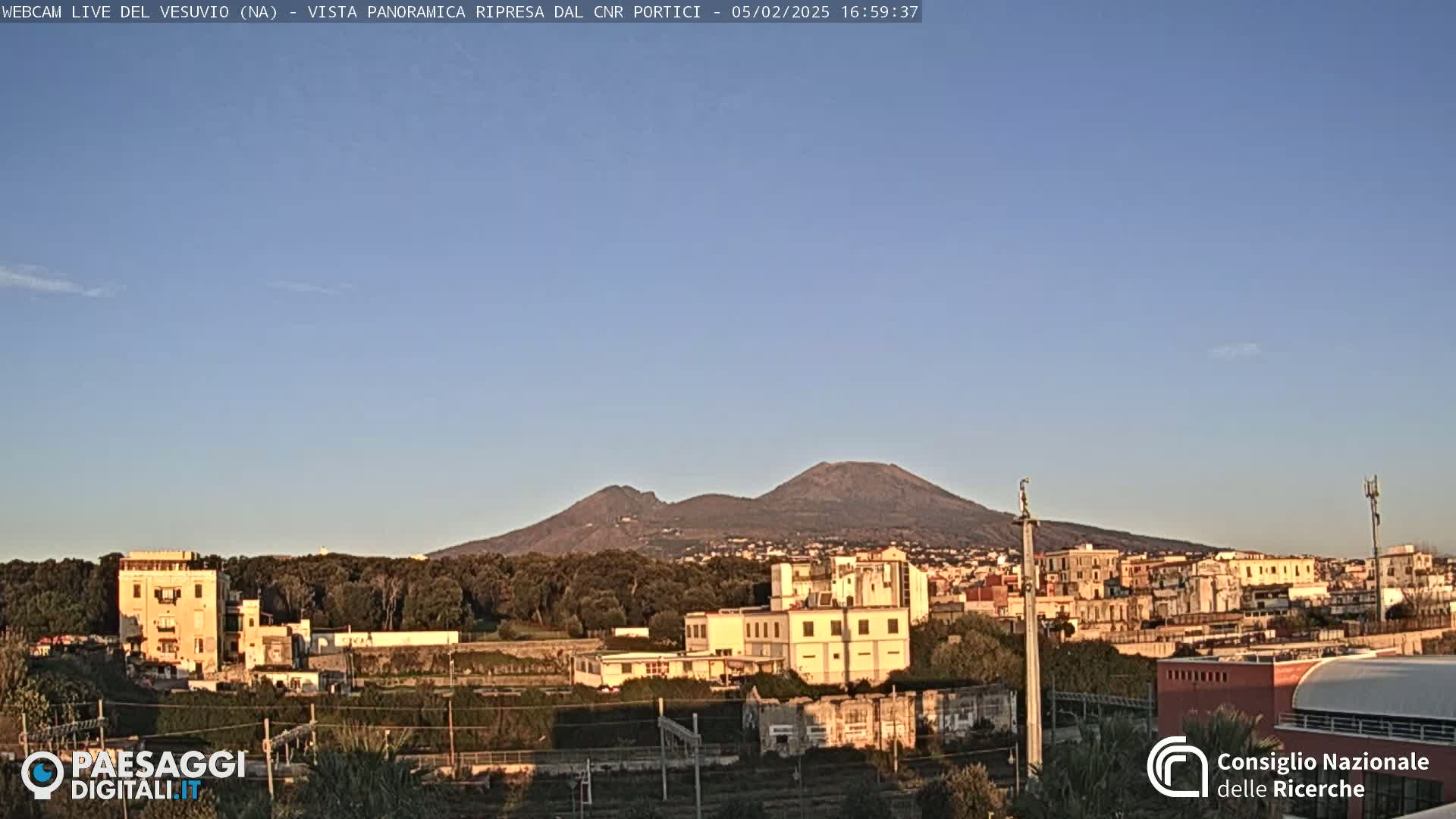 A distant, snow-capped Mount Vesuvius is visible under a clear, light-blue sky above a town with low-rise buildings and sparse vegetation.