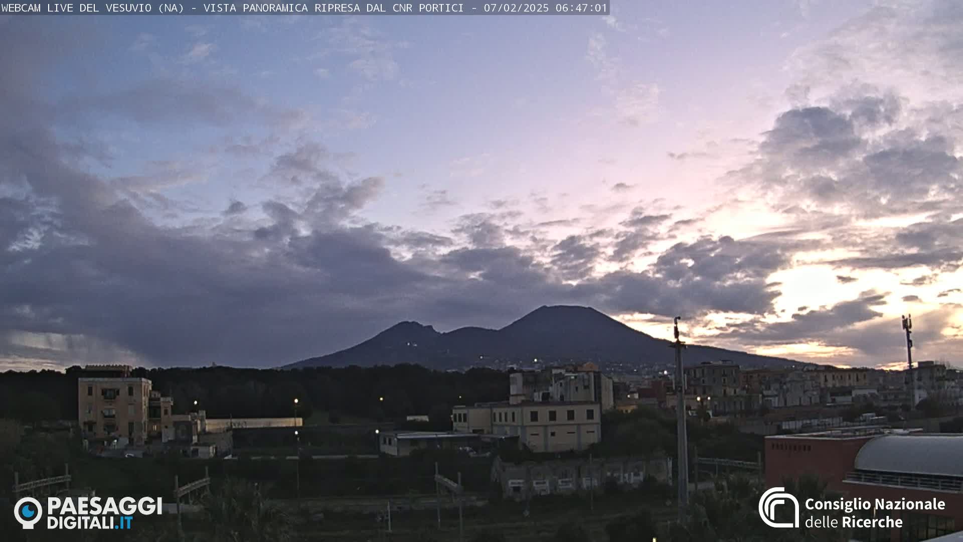 A partly cloudy sunrise illuminates a cityscape with Mount Vesuvius silhouetted in the distance.