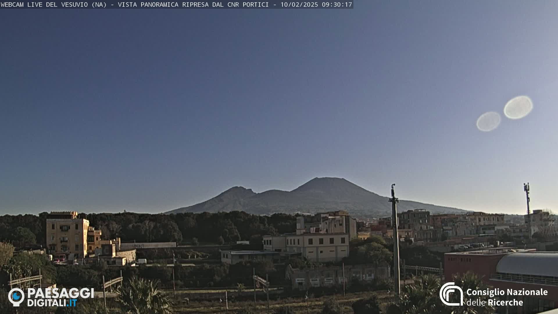 A distant, dark-colored, double-peaked mountain is visible under a clear, light-blue sky above a town with low-rise buildings.