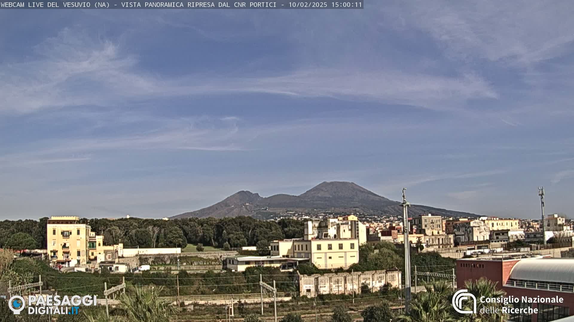 A distant, dark mountain range is visible under a mostly clear, light-blue sky above a flat area of buildings and green vegetation.