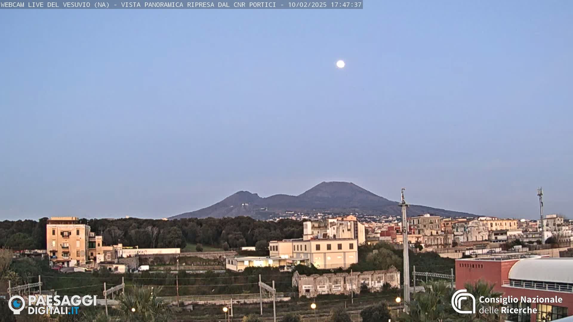 A full moon hangs in a twilight sky above a cityscape and the distant, silhouetted Mount Vesuvius.