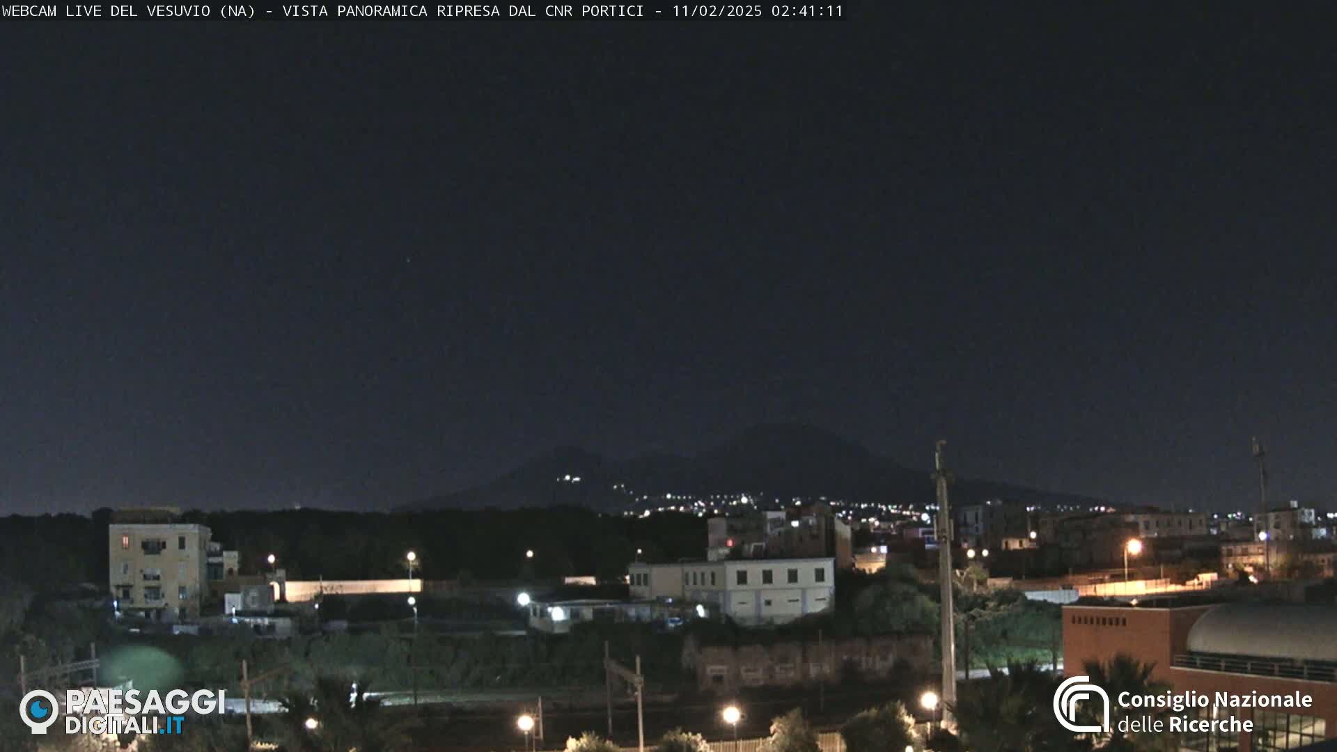 A nighttime view of a city with a distant, dark mountain range illuminated by sparse lights under a clear, dark sky.