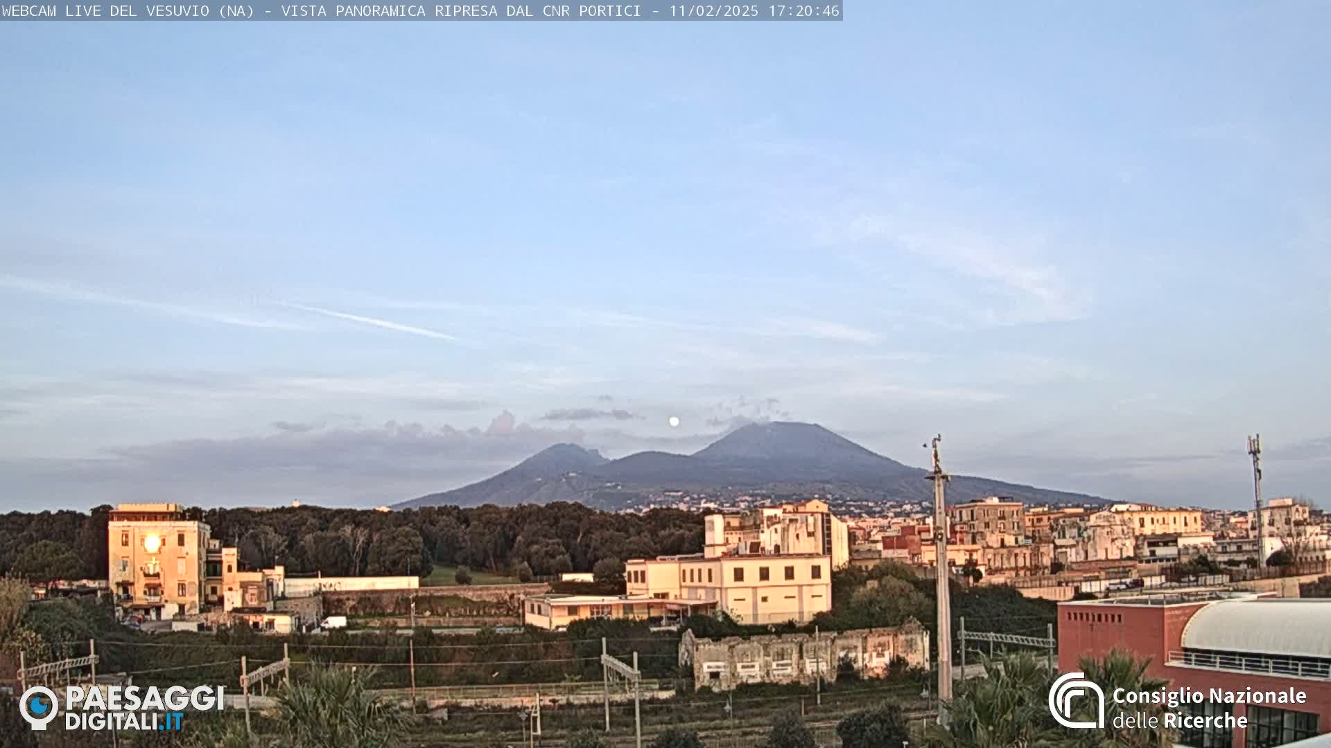A view of Mount Vesuvius in the distance, under a mostly clear sky with a few wispy clouds, overlooking a town with low-rise buildings and some trees.