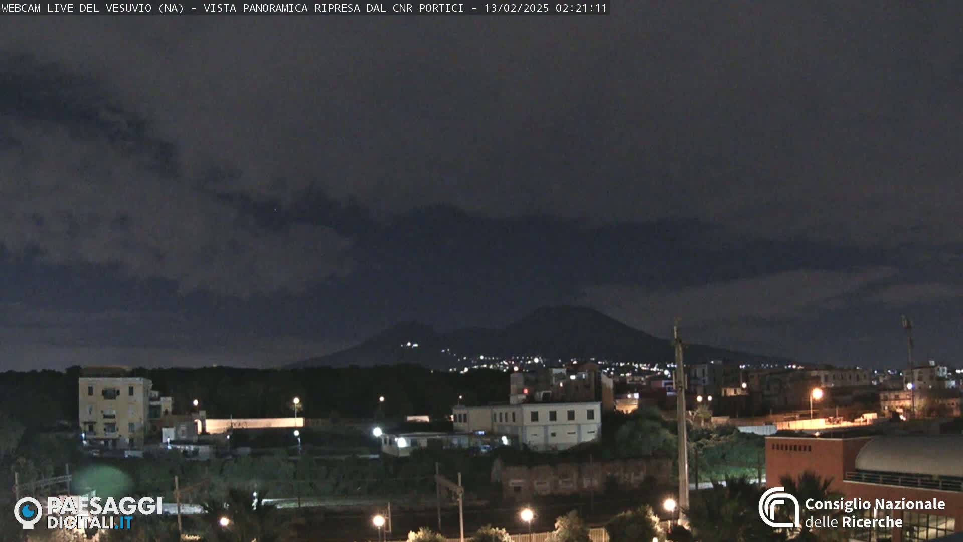 A nighttime view of a city illuminated by lights, with a mountain range faintly visible in the distance under a mostly clear, dark sky.