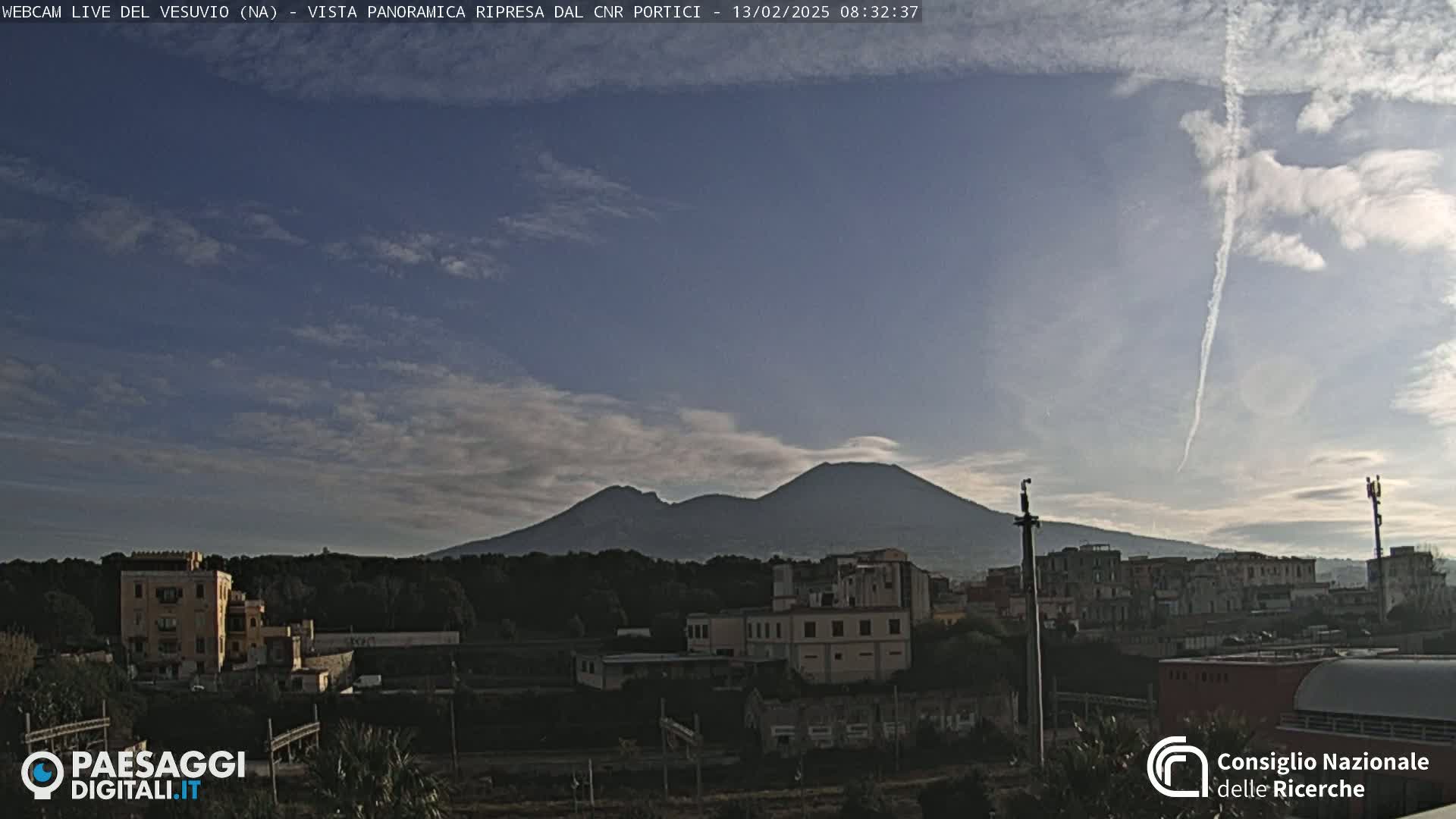 A view of Mount Vesuvius under a mostly cloudy sky with a contrail visible, overlooking a town with buildings and trees.