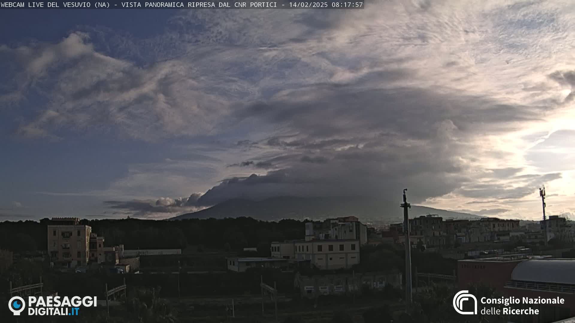 A view of Mount Vesuvius in the distance, partially obscured by a cloudy, overcast sky, with a town and buildings in the foreground.