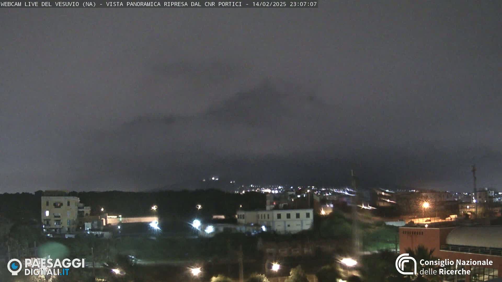 A nighttime view of a city at the foot of a mountain range under a dark, cloudy sky.