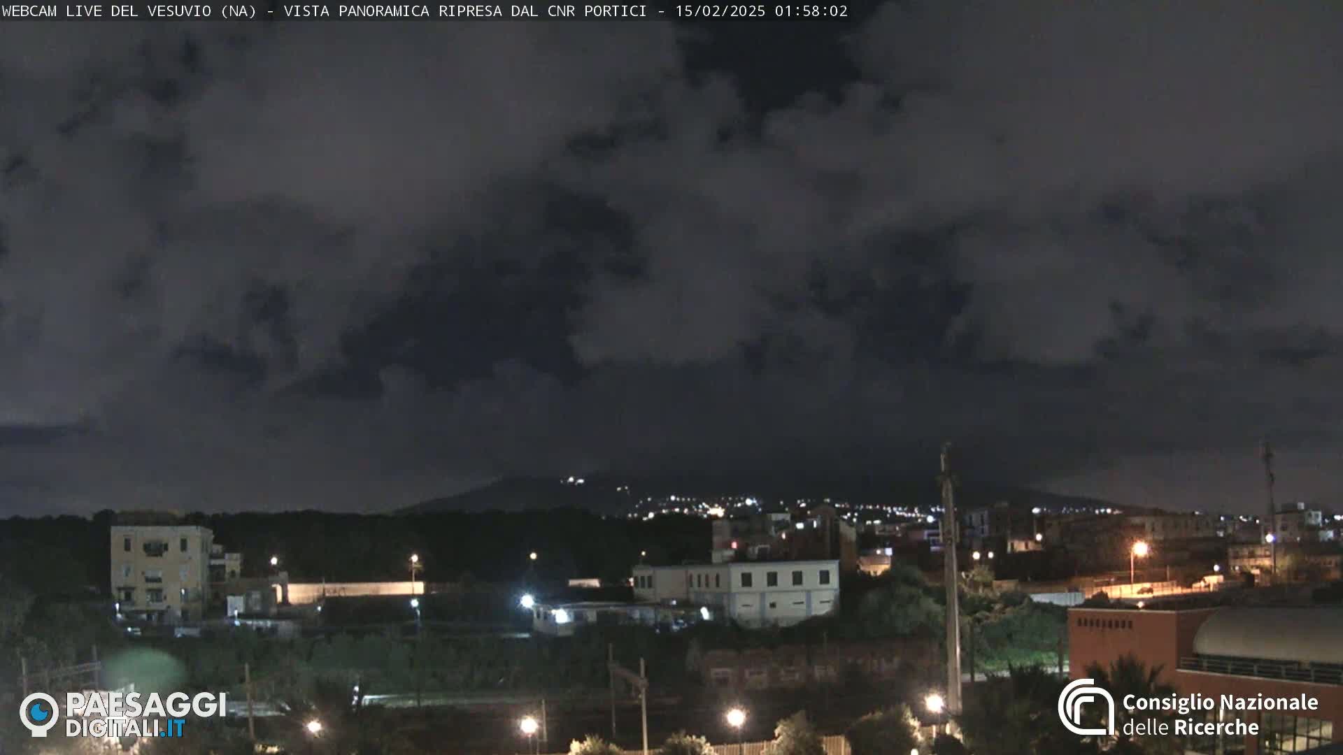 A nighttime panoramic view of a city illuminated by lights under a mostly cloudy sky, with a distant mountain range faintly visible in the background.