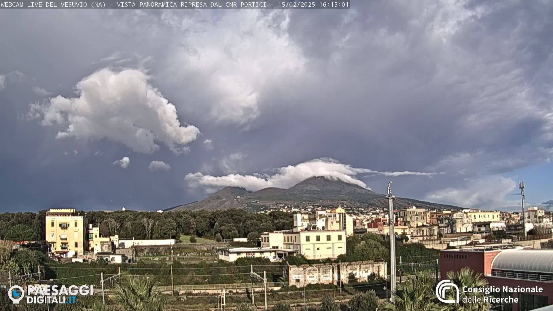 A partly cloudy sky hangs over a town, with a large volcano partially obscured by clouds in the background.