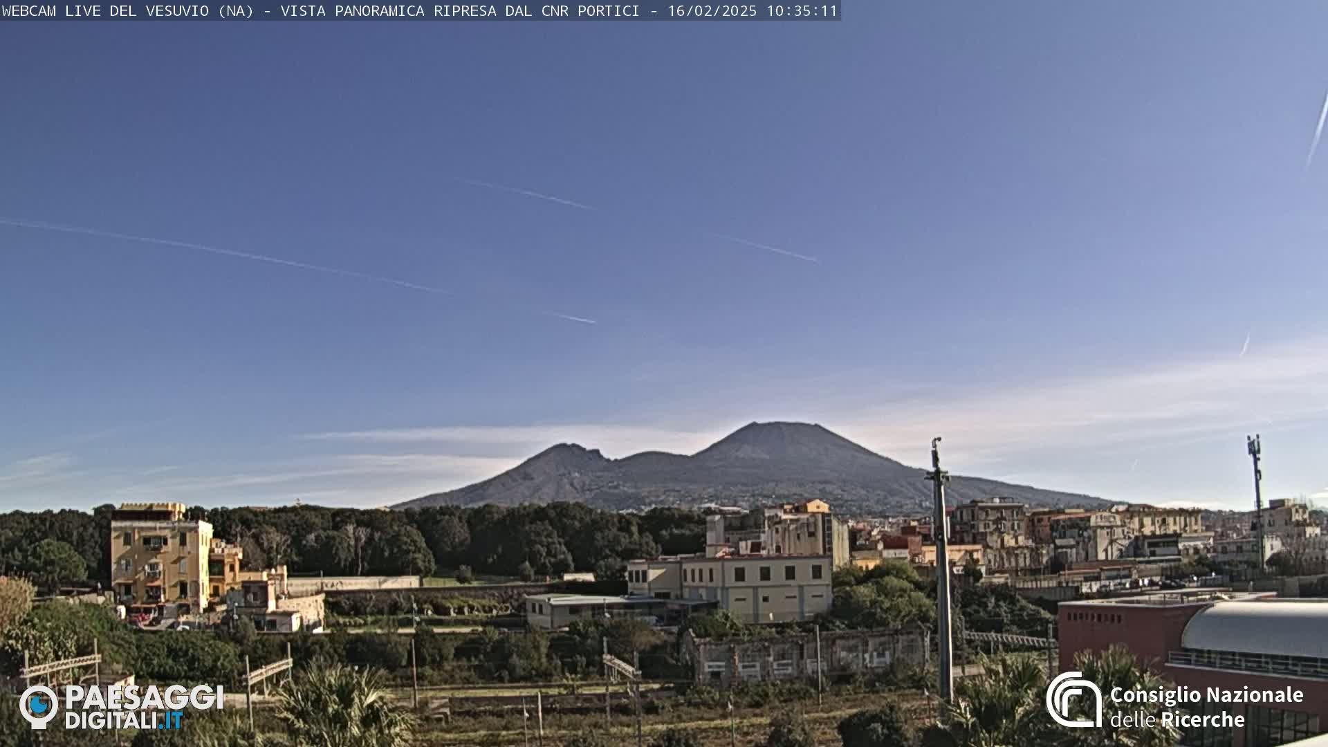 A view of Mount Vesuvius on a clear, sunny day, with buildings and vegetation in the foreground.