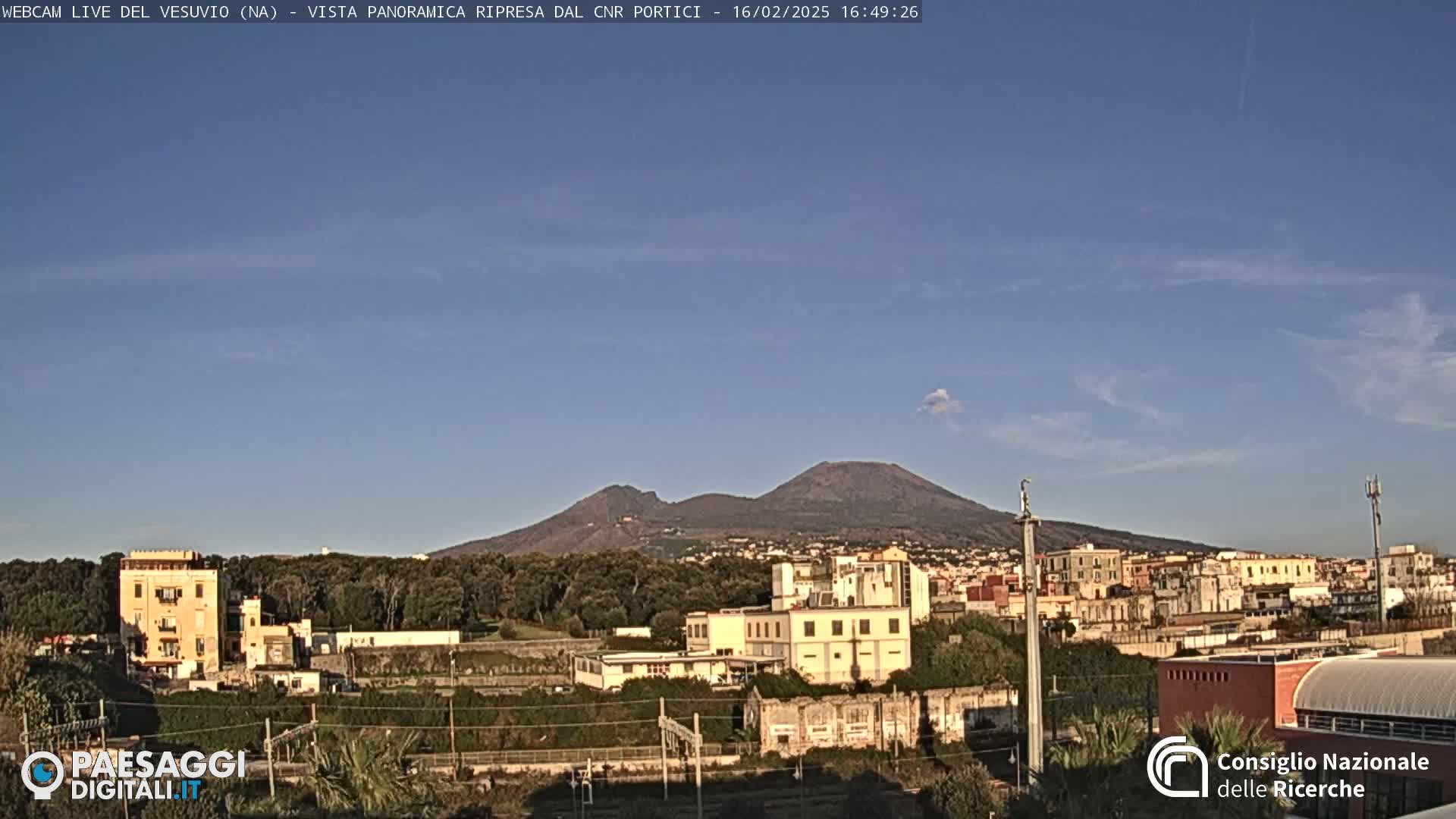 A distant, dark volcano is visible behind a town with low-rise buildings under a mostly clear, light-blue sky.