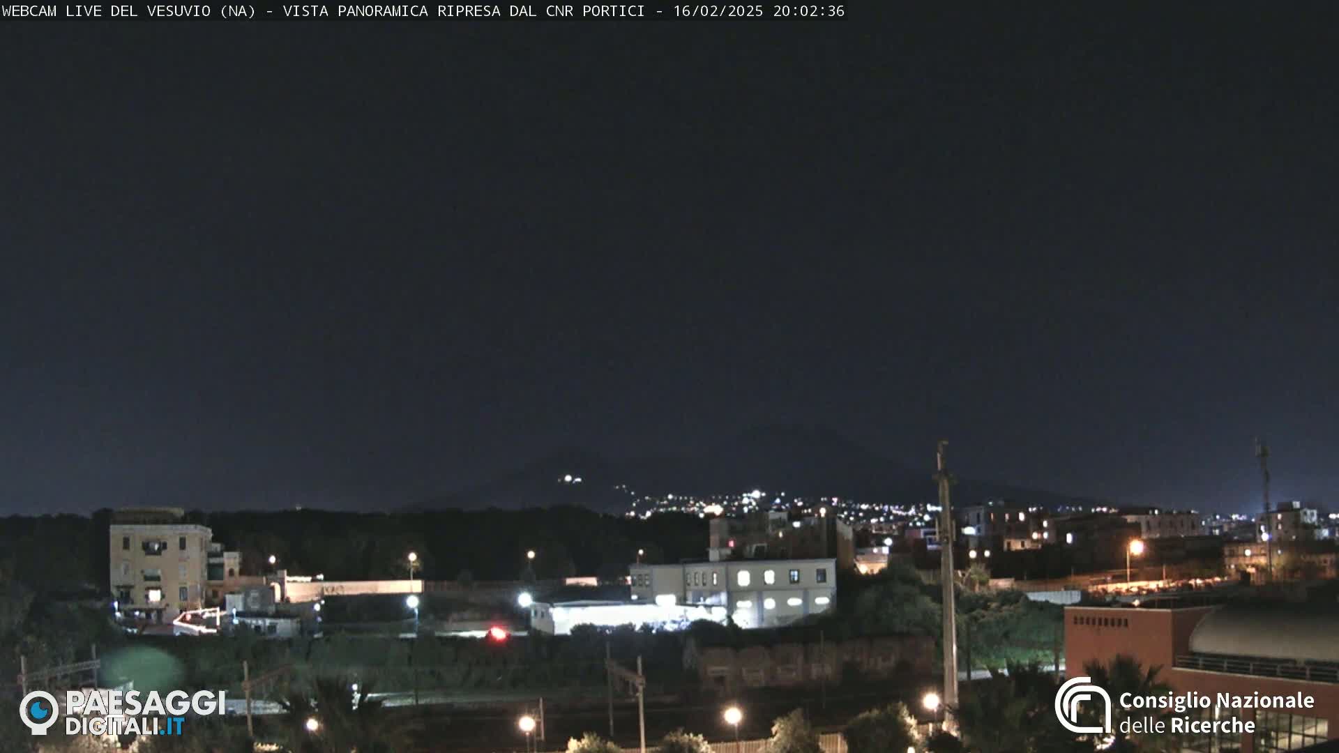 A nighttime, panoramic view of a city with a volcano faintly visible in the distance under a dark sky.