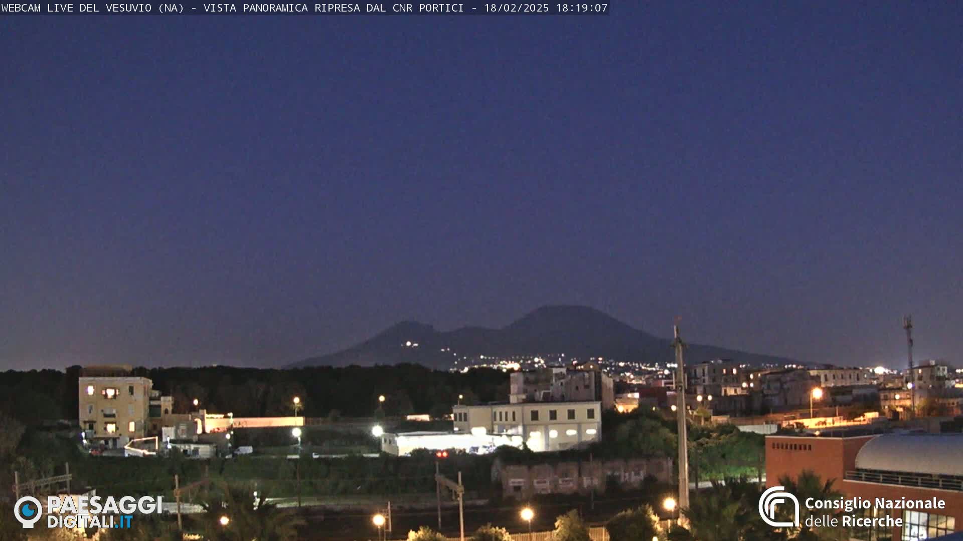 A nighttime view of a city with Mount Vesuvius in the background under a clear, dark blue sky.