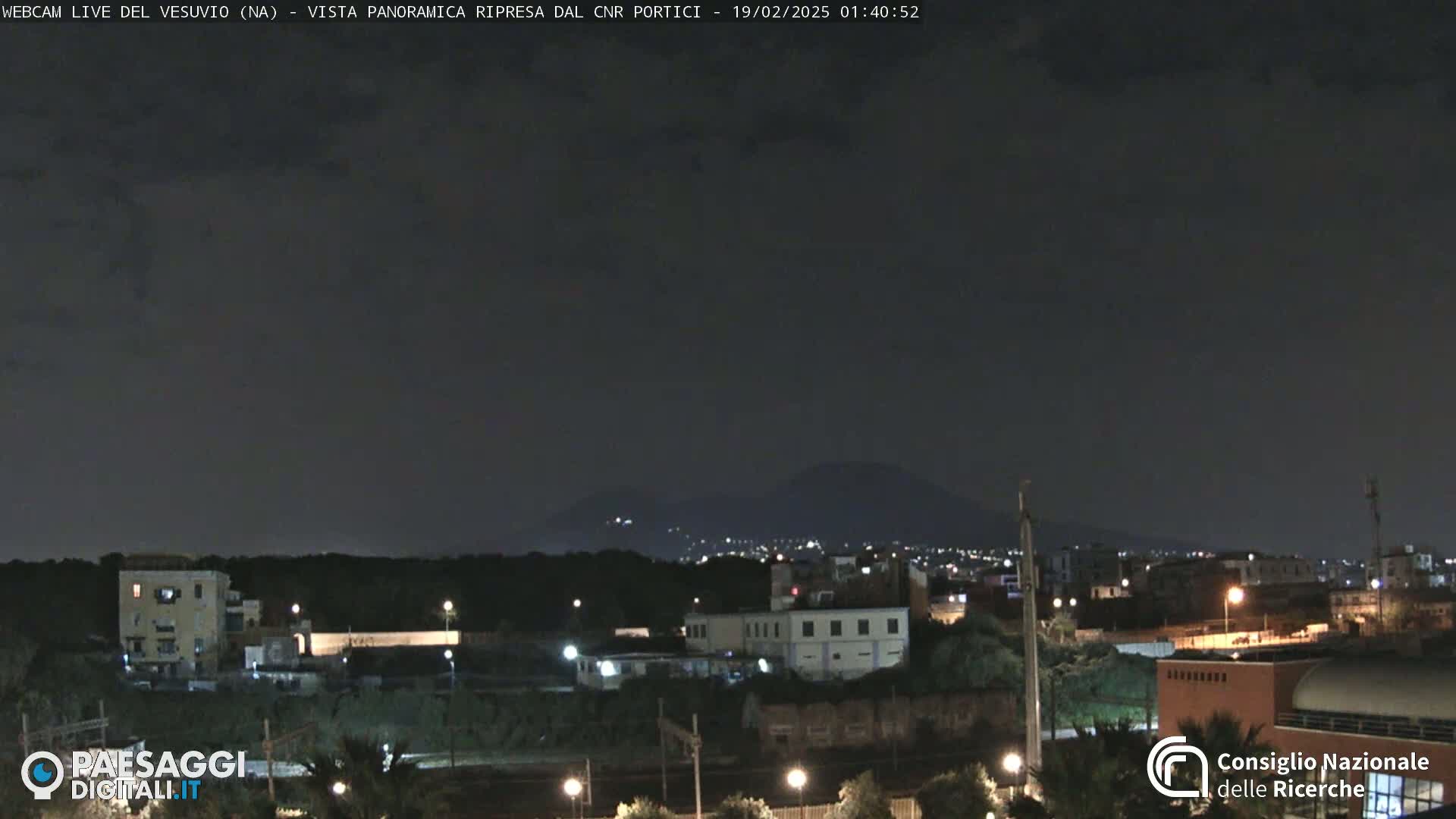 A nighttime view of a city with a dark, overcast sky and a mountain faintly visible in the distance.