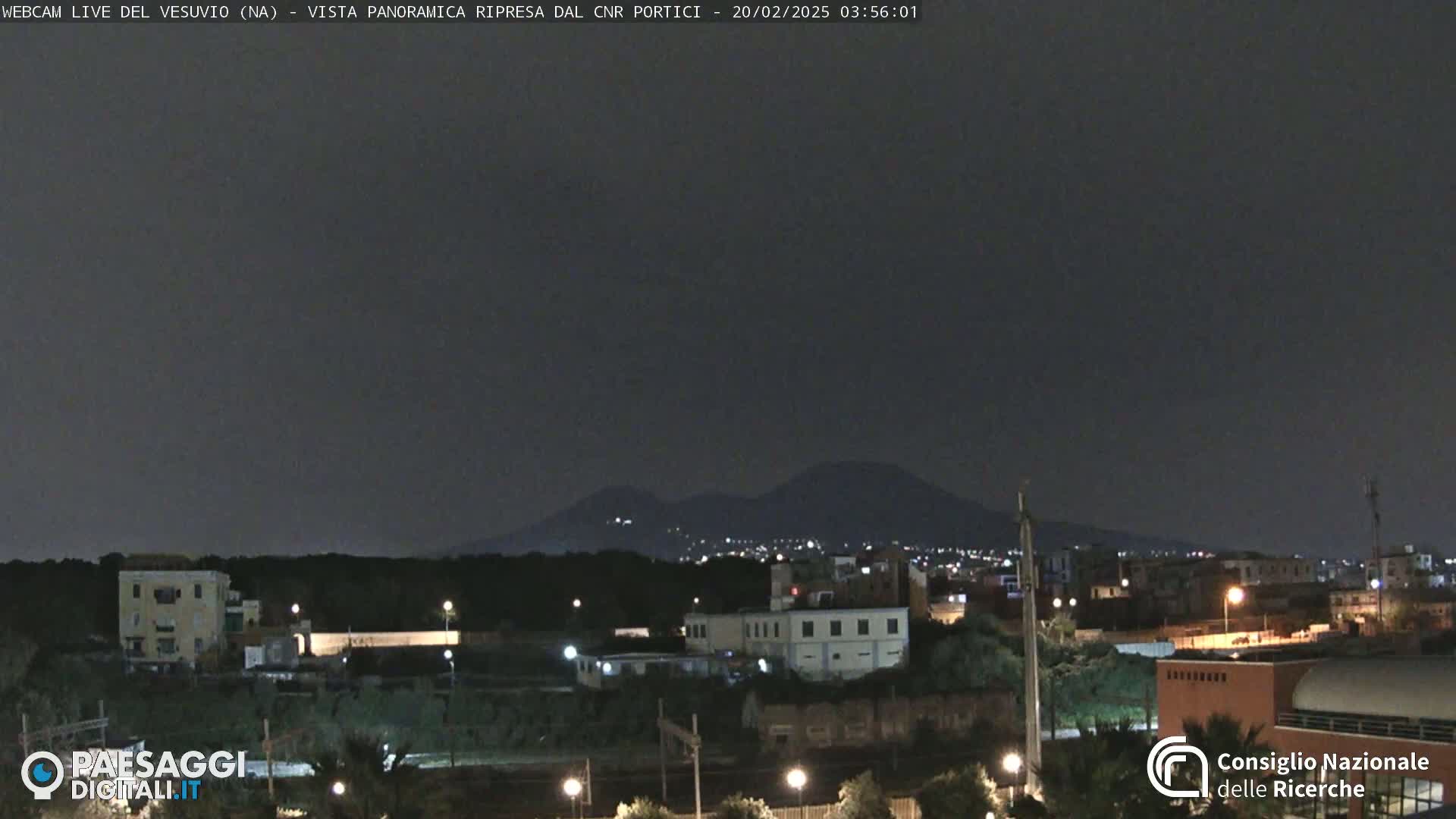 A nighttime, long shot of a city with a mountain range in the distance under a dark sky.