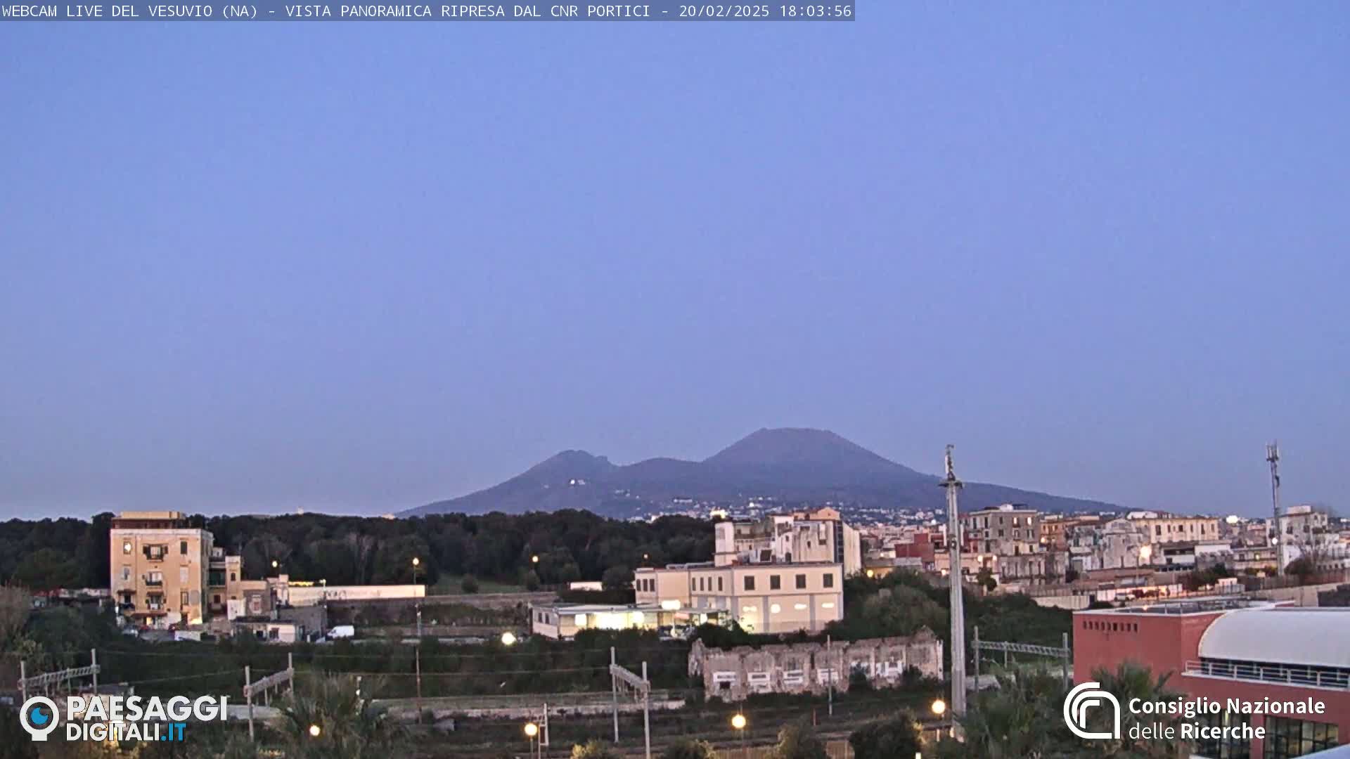 A distant, dark-colored mountain range is visible in the dusky evening light above a cityscape of low-rise buildings and train tracks.