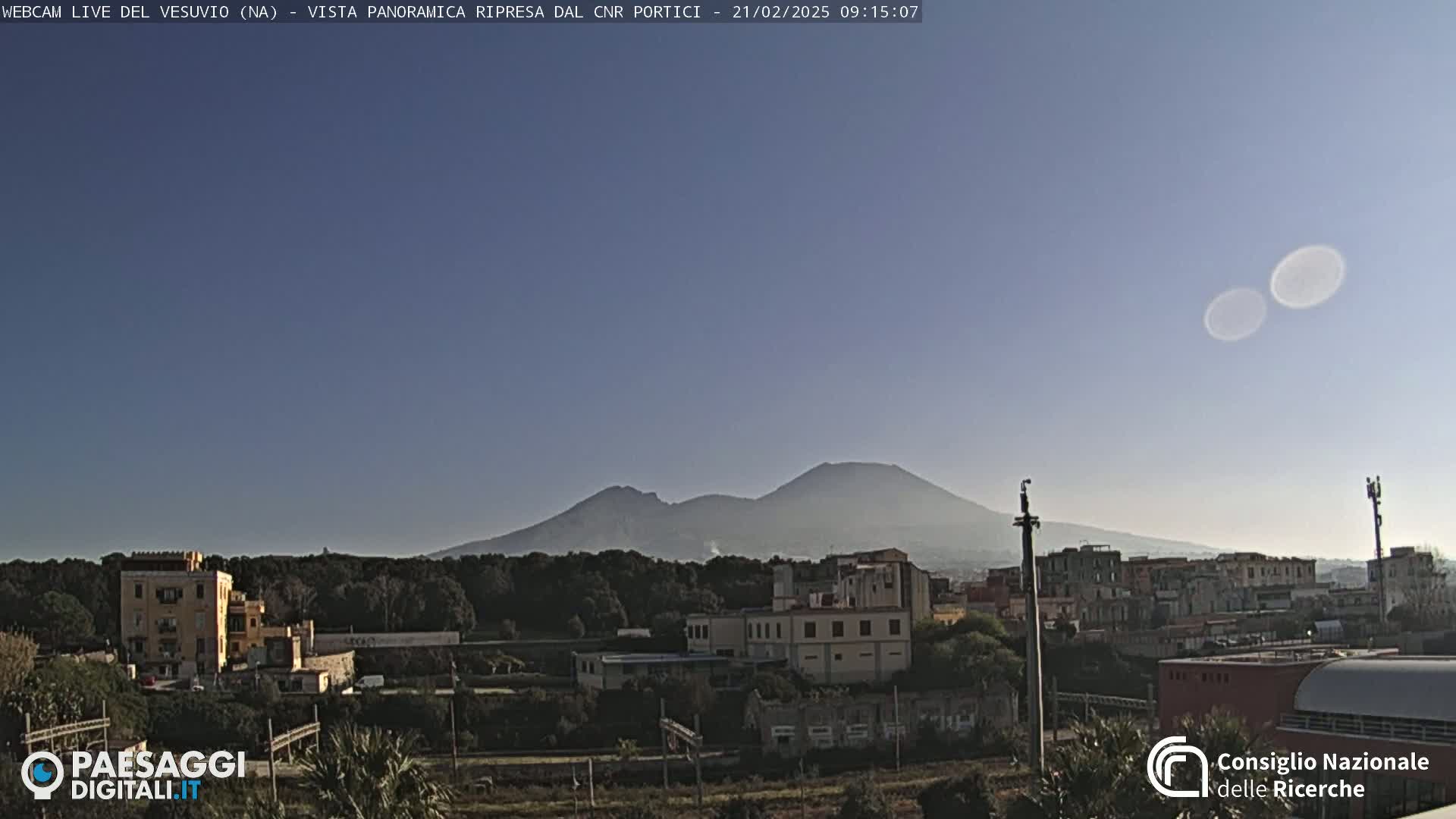 A distant, hazy view of Mount Vesuvius under a clear, light-blue sky, overlooking a town with buildings and trees in the foreground.