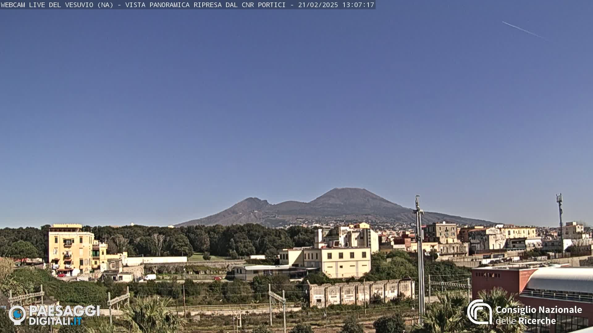 A clear, sunny day shows Mount Vesuvius in the distance behind a town with low-rise buildings and trees.