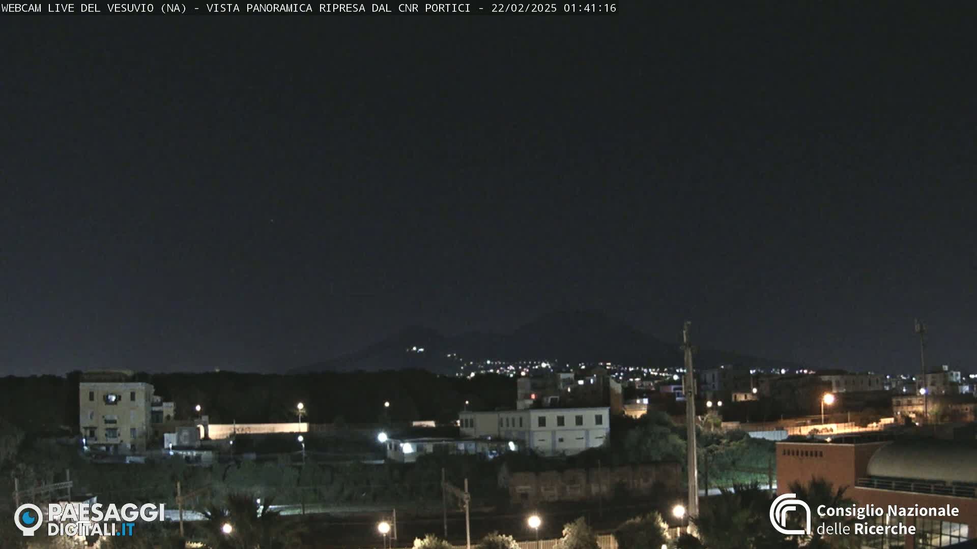 A nighttime, panoramic view of a city illuminated by streetlights with a dark mountain range in the background under a clear sky.