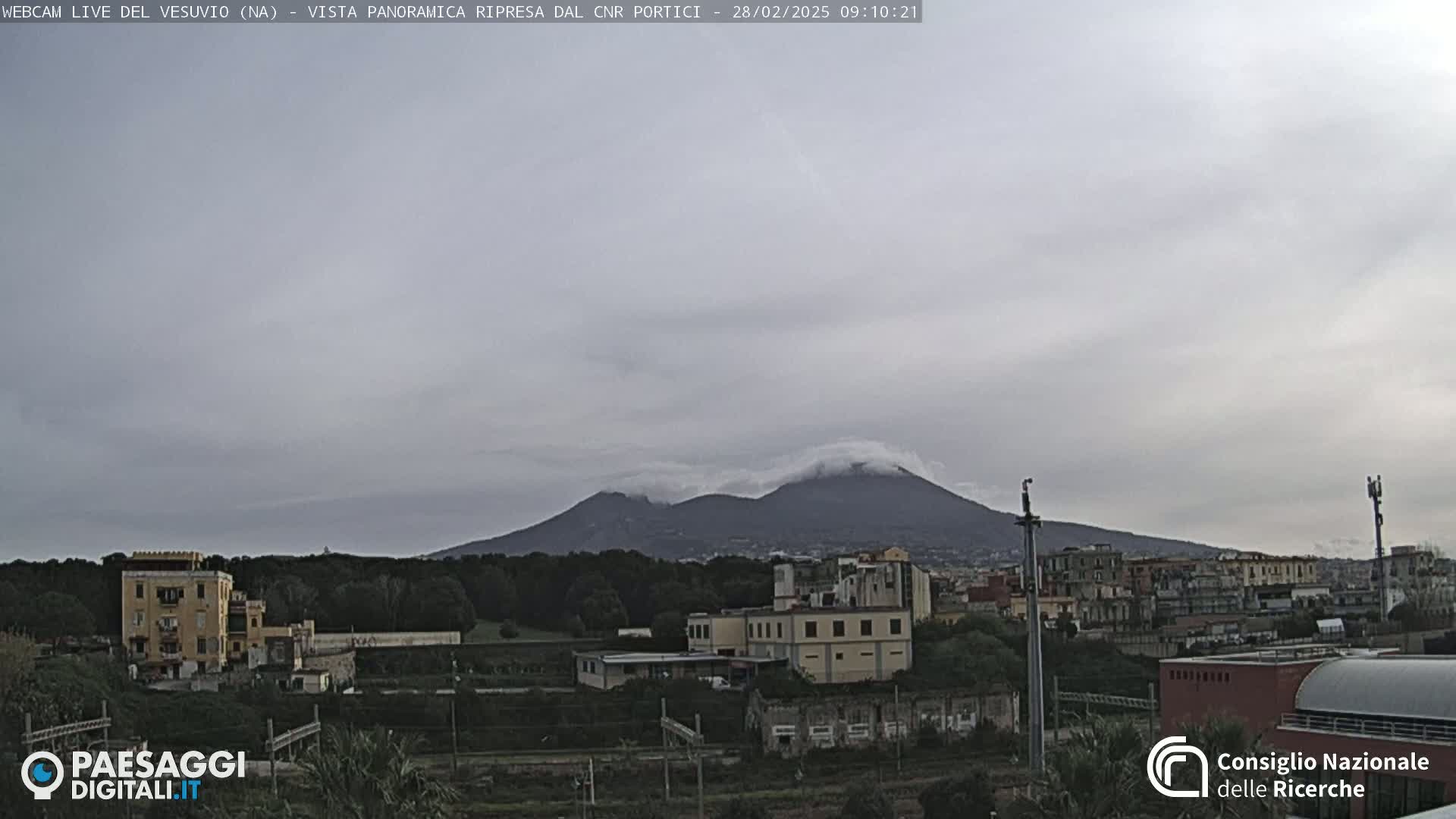Mount Vesuvius is visible in the distance, partially obscured by clouds, over a cityscape under an overcast sky.