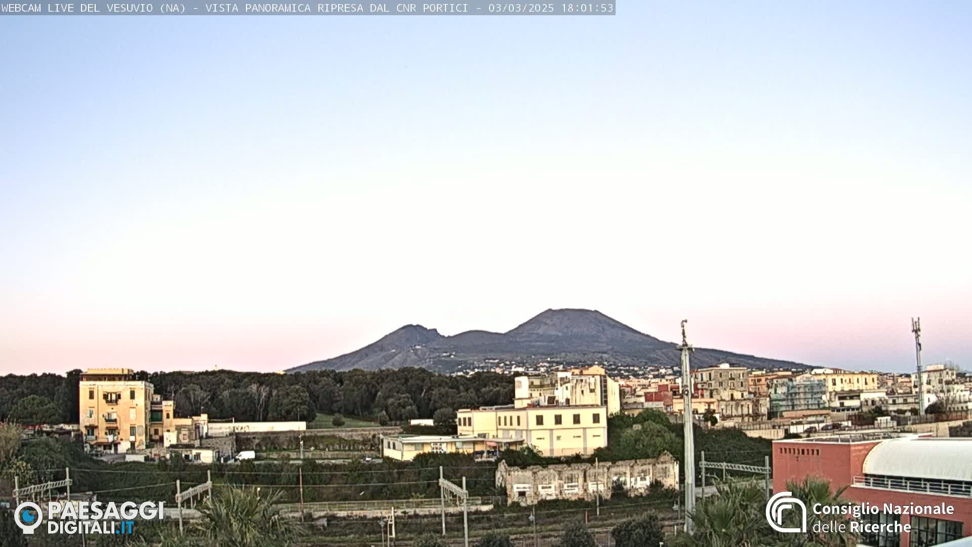 A distant, dark mountain range is visible under a pale pink and light blue twilight sky above a city of low-lying buildings and train tracks.