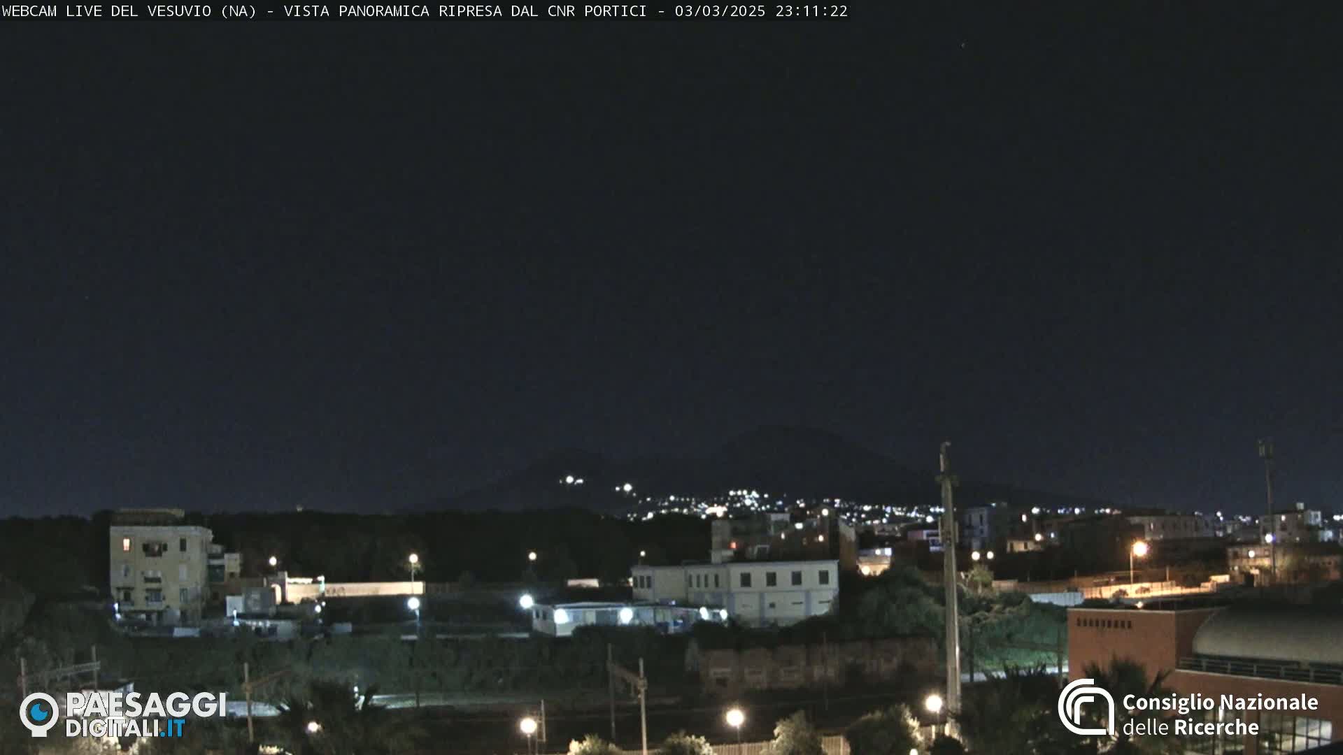 A nighttime view of a town illuminated by streetlights, with a dark mountain range in the background under a clear, dark sky.