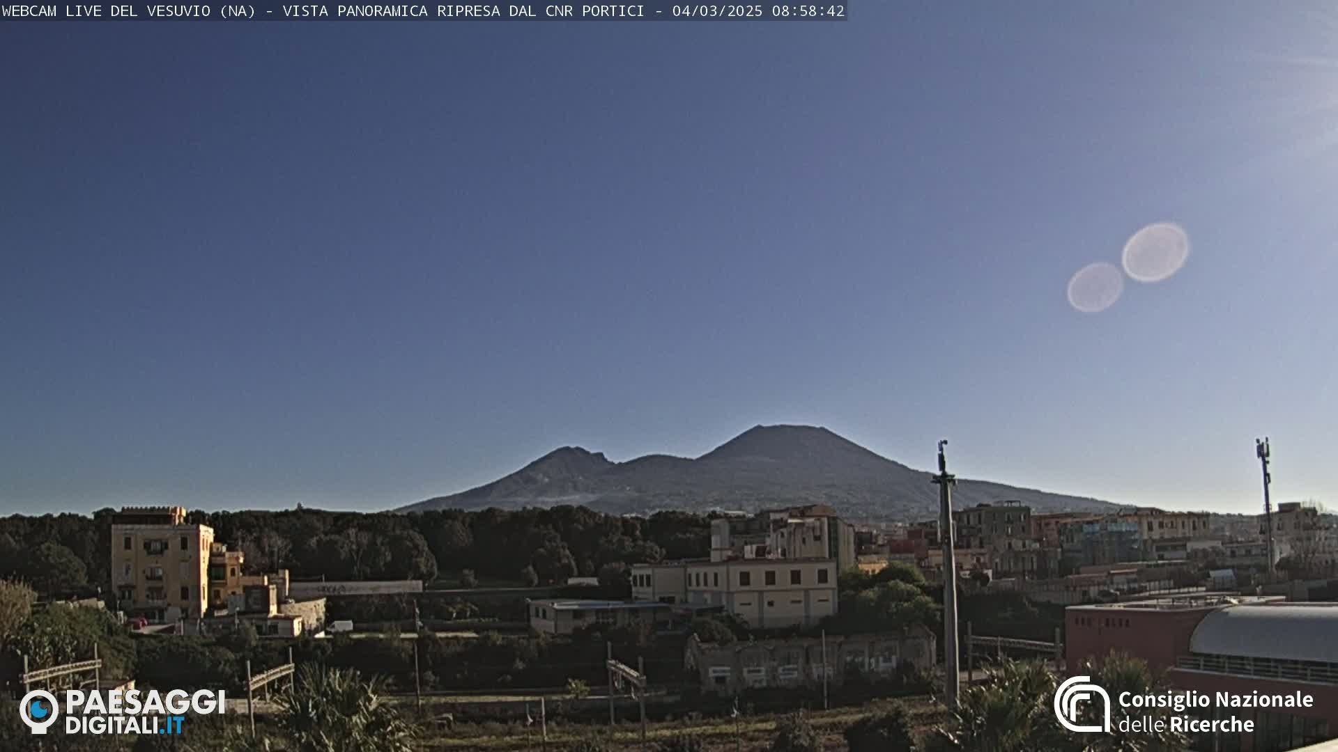 A long shot of Mount Vesuvius on a clear, sunny day, with buildings and vegetation in the foreground.