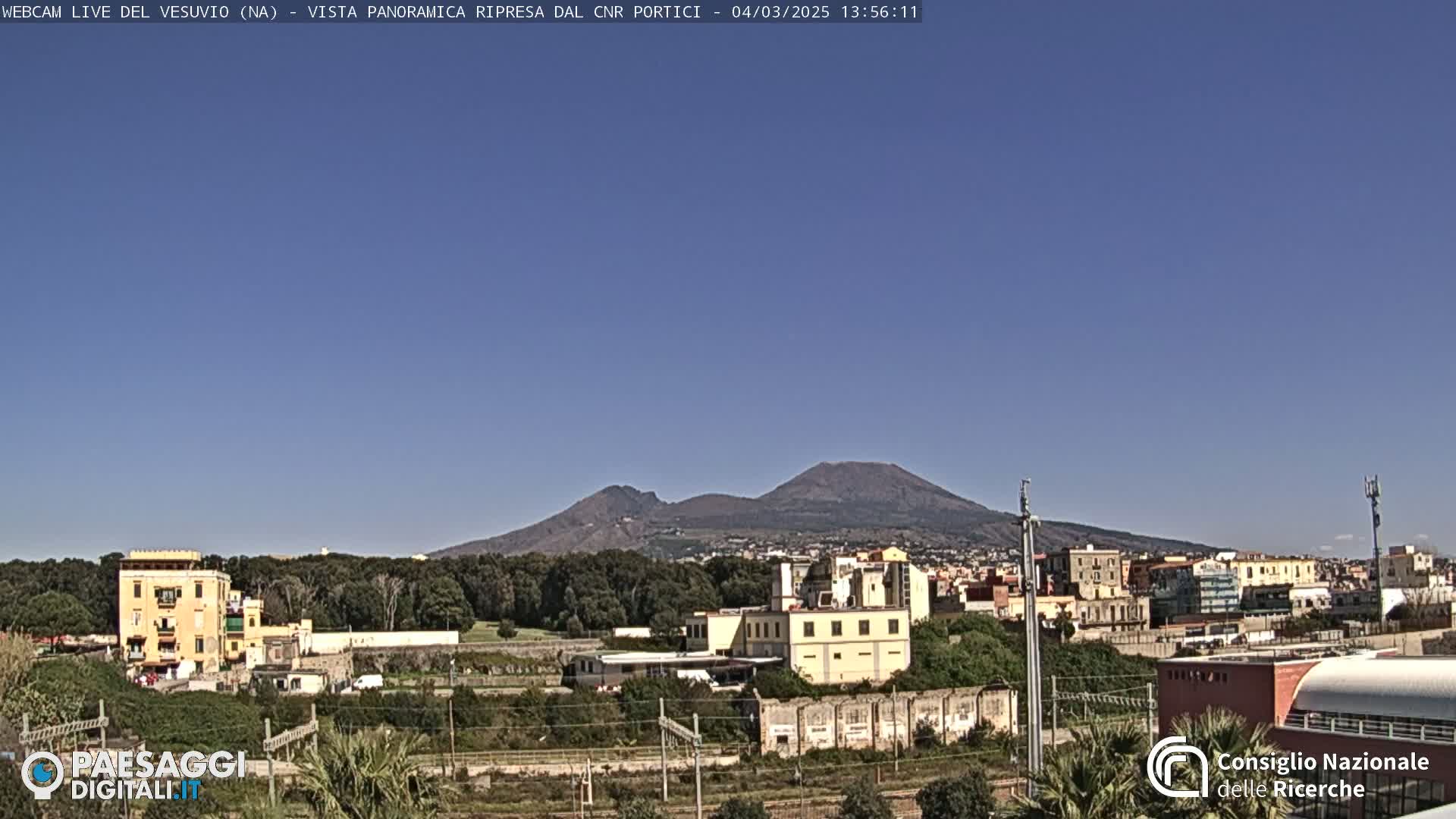 A distant, dark volcano is visible under a clear blue sky above a cityscape of low buildings and green vegetation.