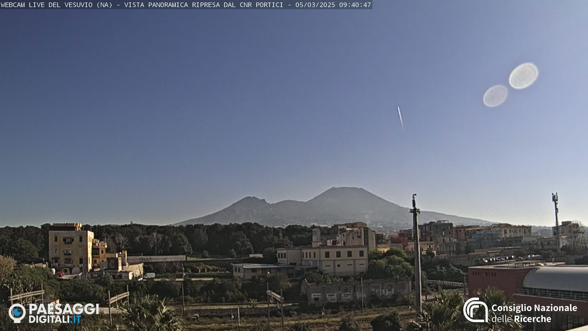 Mount Vesuvius is visible in the distance on a clear, sunny day, behind a town with low buildings and trees.