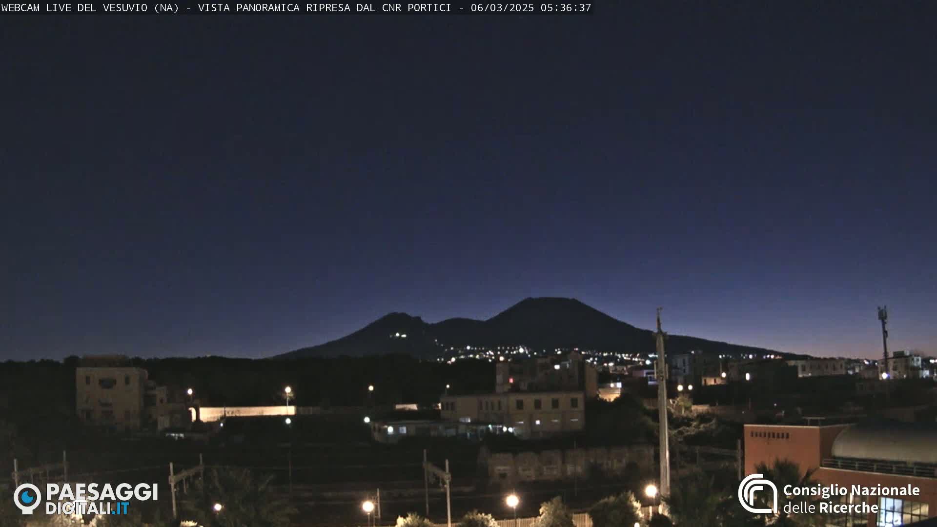 A nighttime view of a city with a dark, mountainous silhouette in the background under a clear, dark-blue sky.
