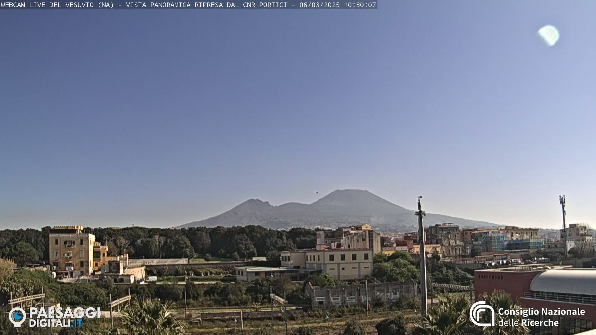 A distant, hazy view of Mount Vesuvius under a clear, bright sky, overlooking a cityscape with low-rise buildings and vegetation.