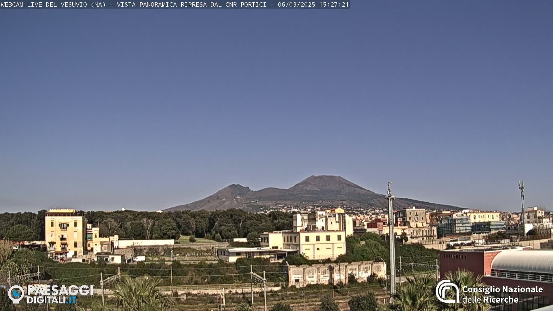 A view of Mount Vesuvius in the distance, under a clear blue sky, with buildings and trees in the foreground.
