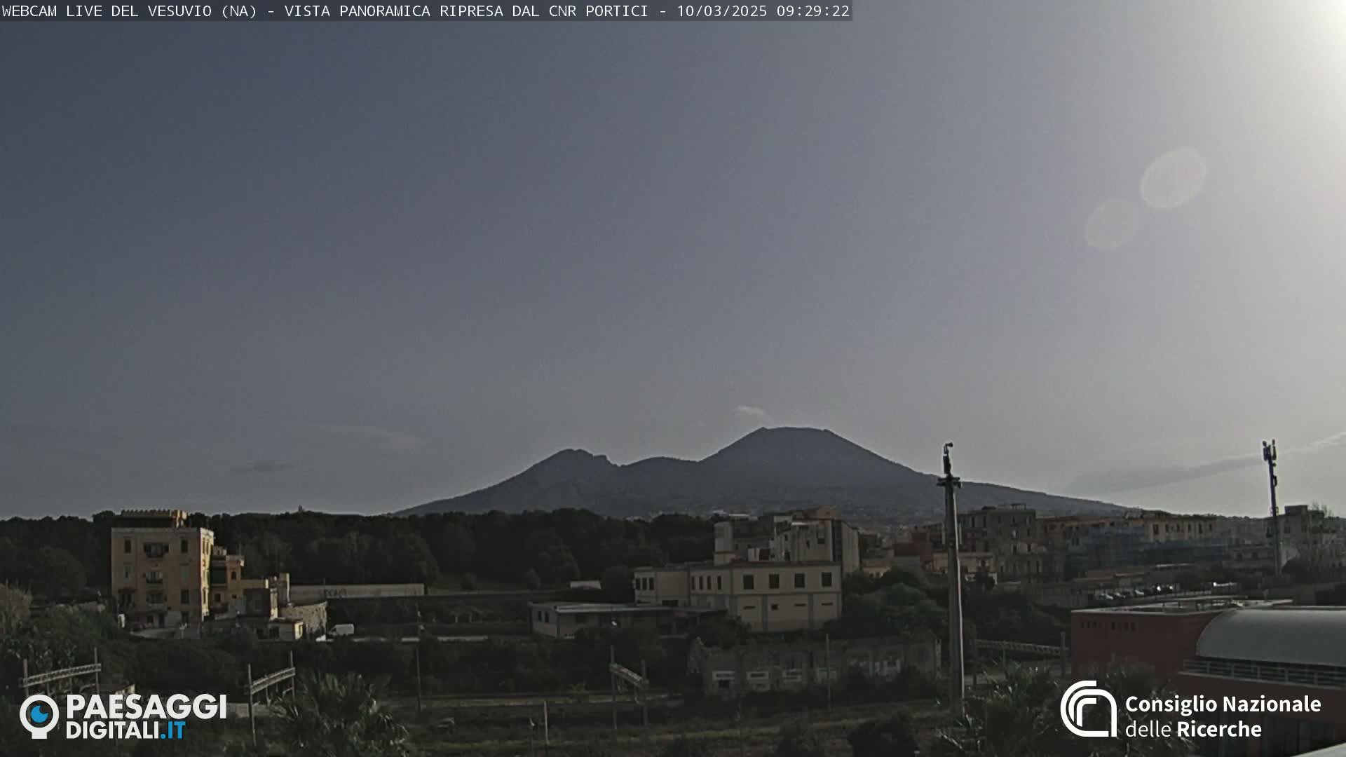 A distant, dark volcano is visible under a hazy, light-grey sky above a town with low buildings and trees.