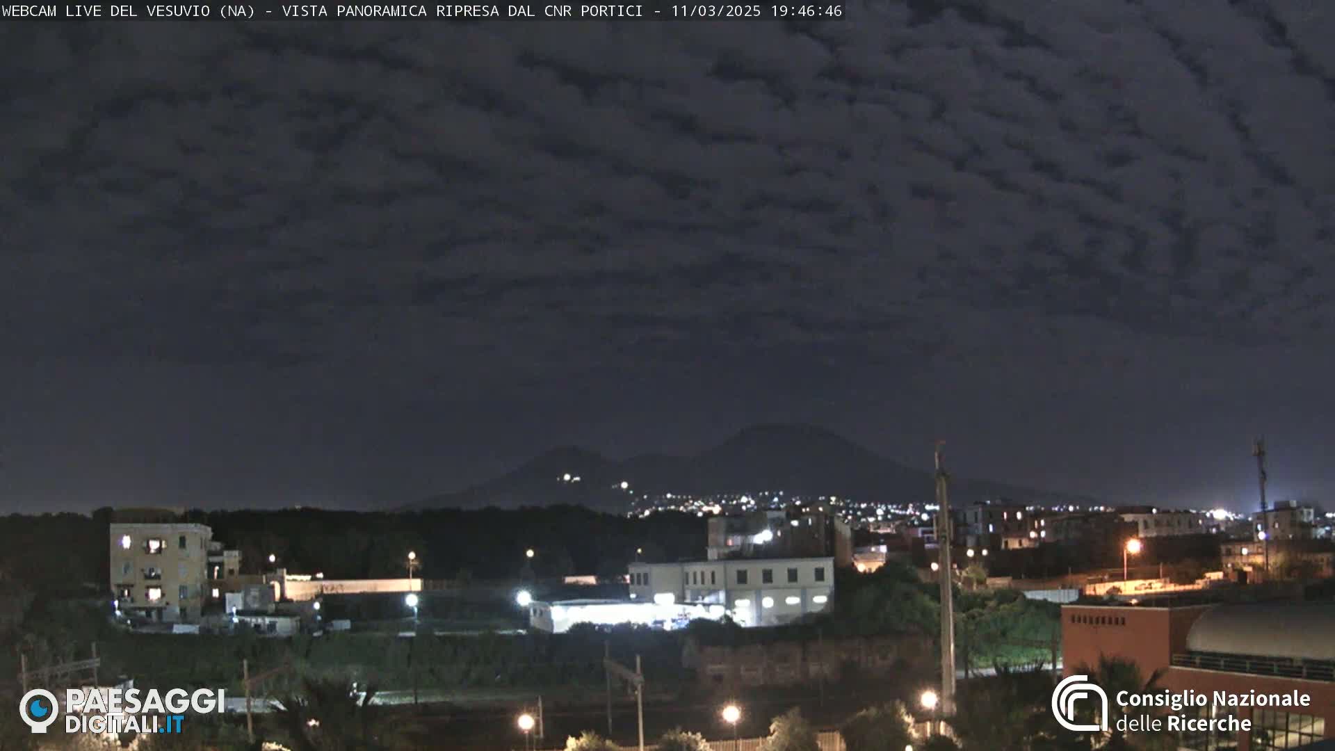 A nighttime view of a city illuminated under a mostly cloudy sky, with two distant mountains visible on the horizon.