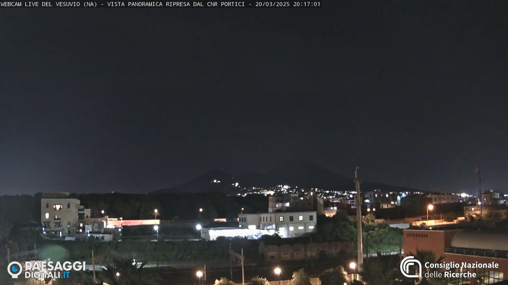A nighttime view of a town illuminated by streetlights, with a dark mountain silhouetted in the distance under a clear, dark sky.