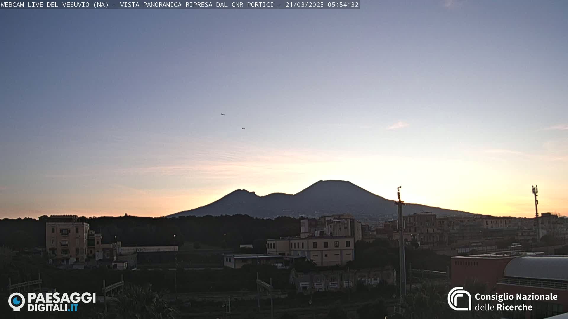 A distant, dark mountain range is silhouetted against a pale sunrise sky, with several buildings and some trees in the foreground.