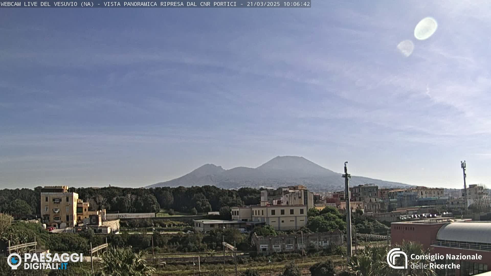 A view of Mount Vesuvius in the distance under a mostly clear, light blue sky, with buildings and trees in the foreground.