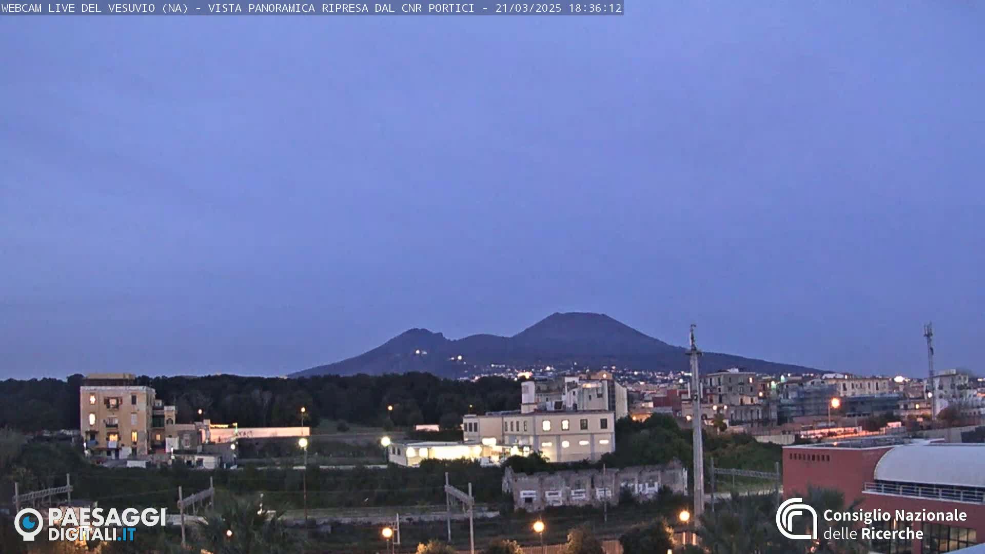 A twilight view shows Mount Vesuvius in the distance, overlooking a cityscape with industrial buildings and residential areas under a clear, dusky sky.