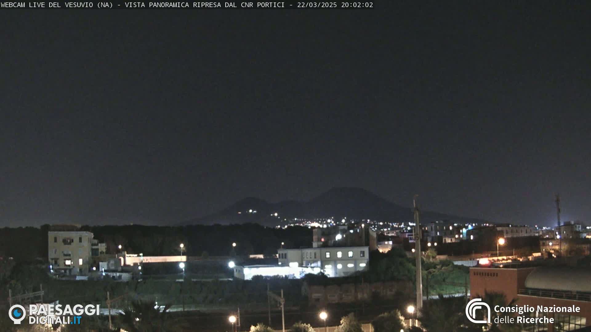 A nighttime long shot of a city illuminated by lights with a mountain range in the background under a clear, dark sky.
