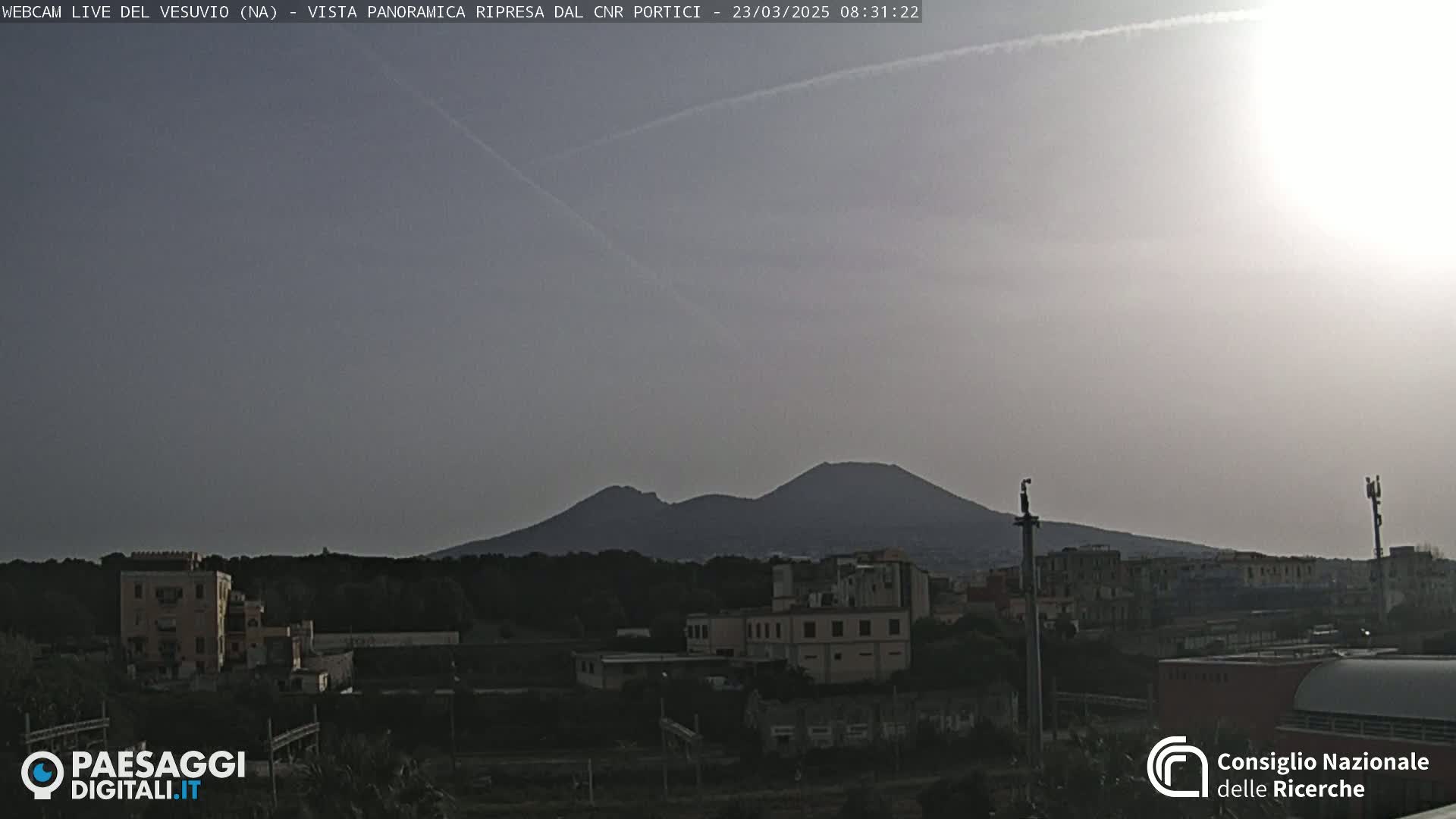 A hazy, bright day reveals a distant, dark silhouette of Mount Vesuvius overlooking a cityscape.