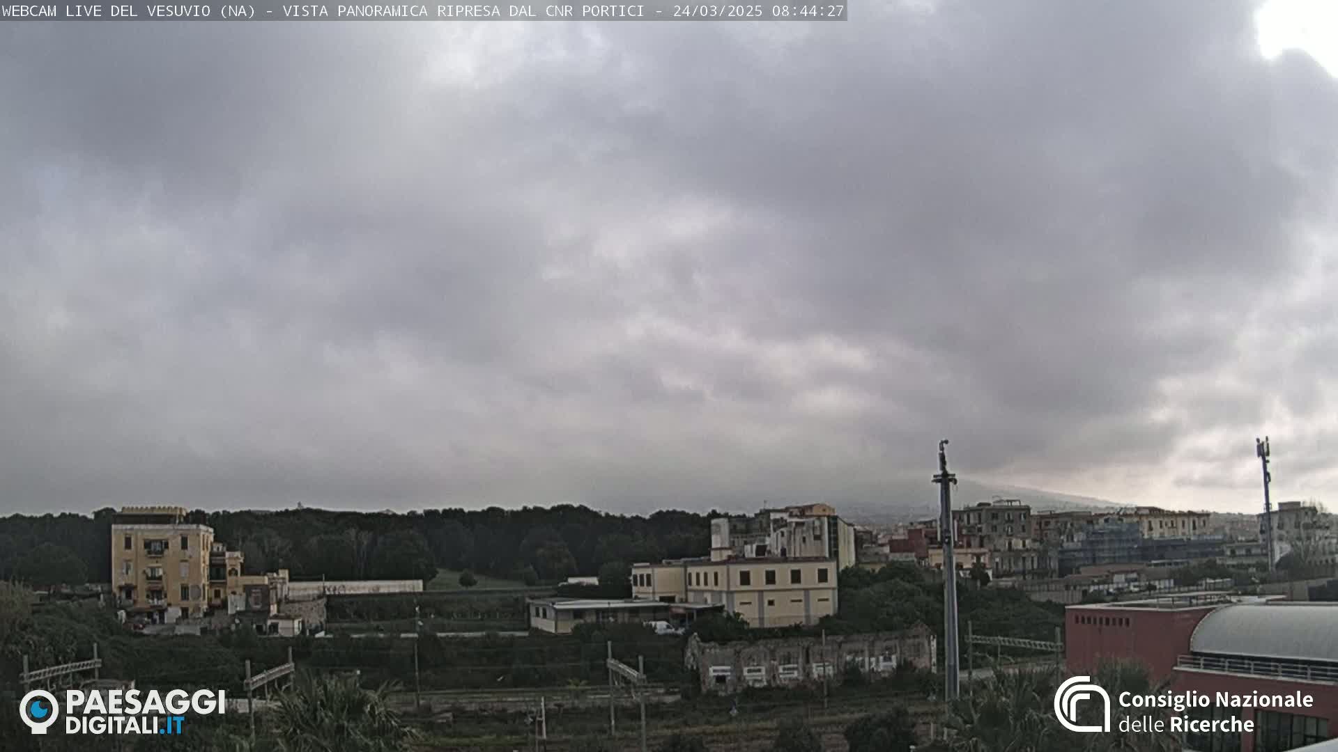 A view of a town under a cloudy sky, showing buildings and trees, with a distant mountain visible.