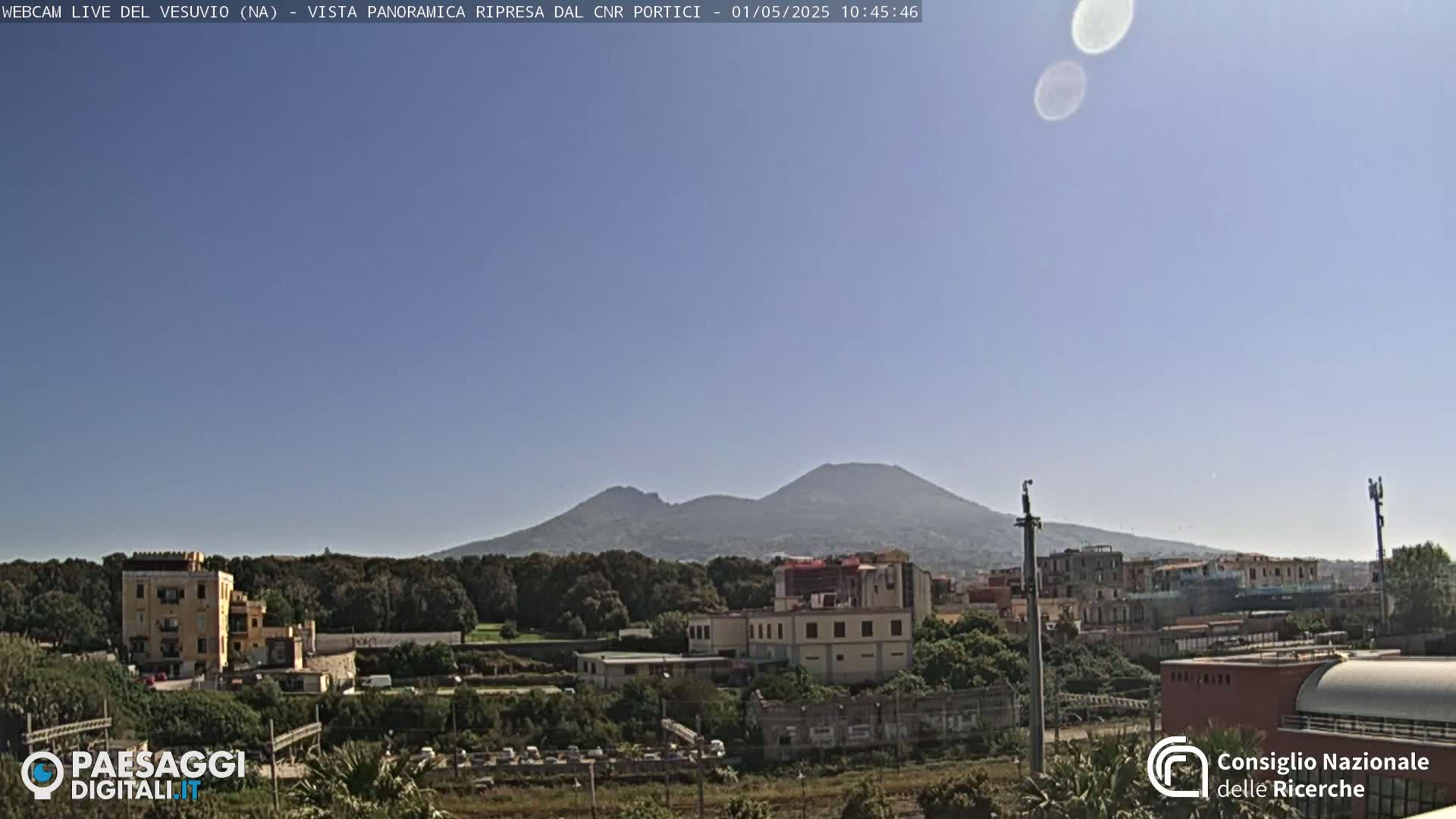 A distant, hazy view of Mount Vesuvius under a clear, sunny sky, overlooking a cityscape with buildings and greenery in the foreground.