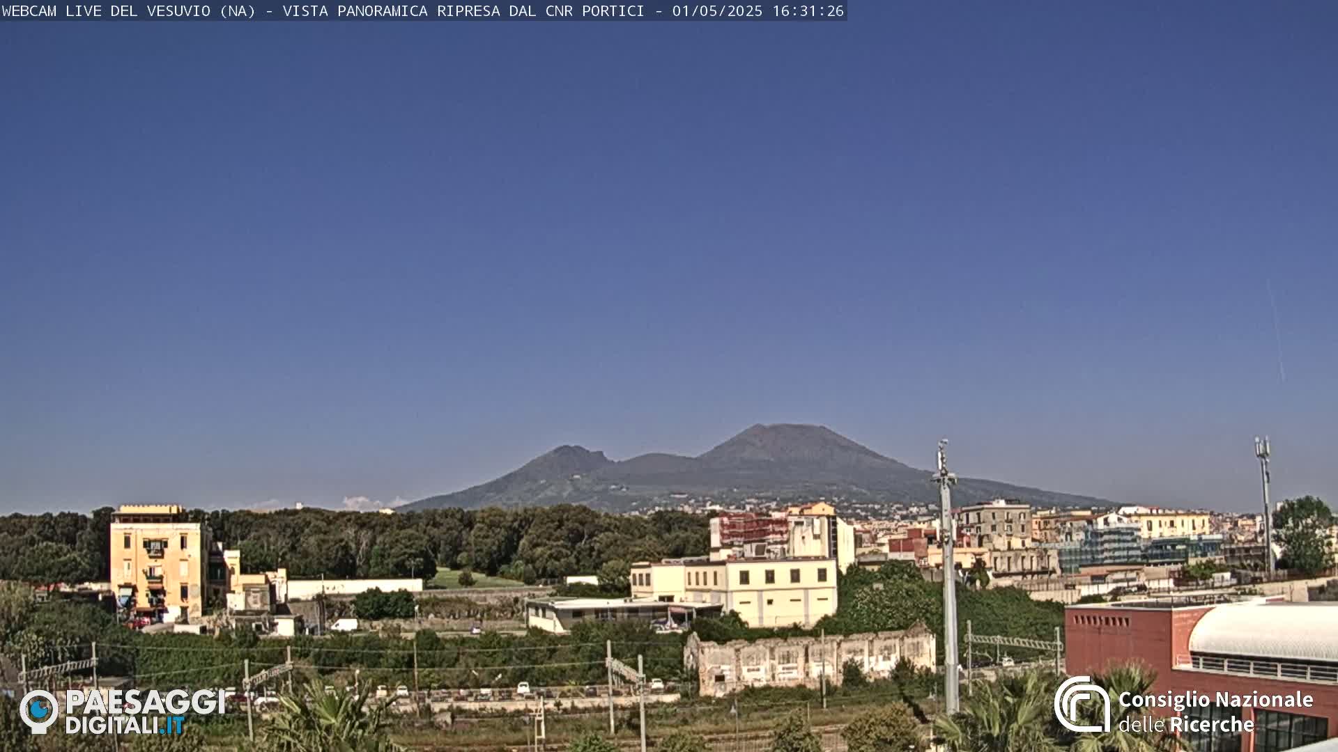 A view of Mount Vesuvius in the distance, under a clear blue sky, overlooking a town with buildings and greenery.