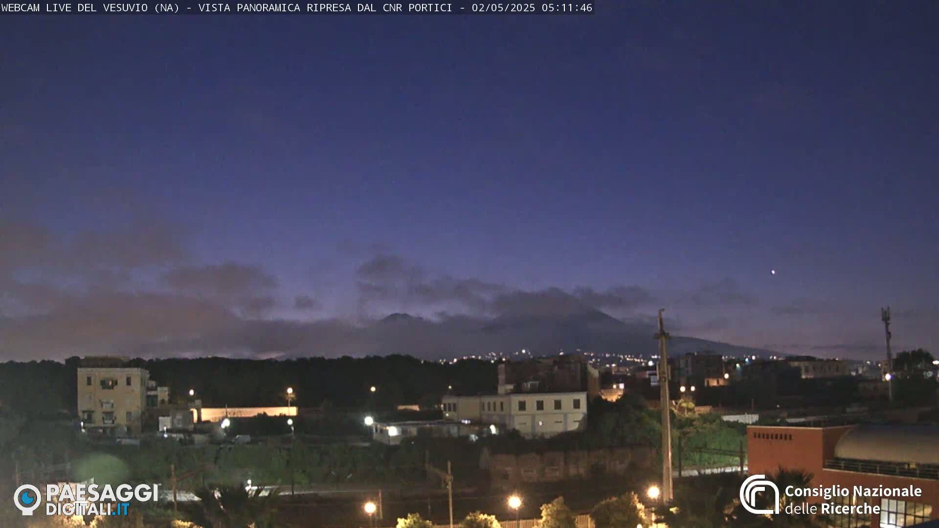 A nighttime view of a city with a volcano in the distance under a mostly clear, dark-blue sky.