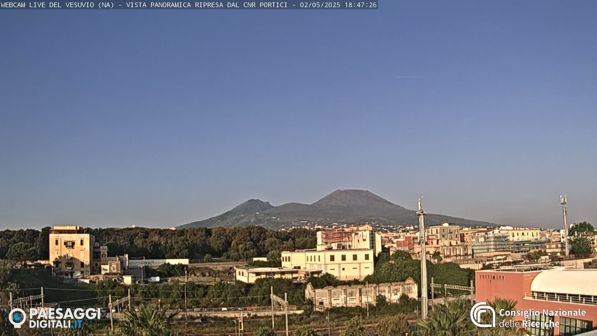 A distant, large volcano is visible over a flat landscape of buildings and trees under a clear, blue sky.