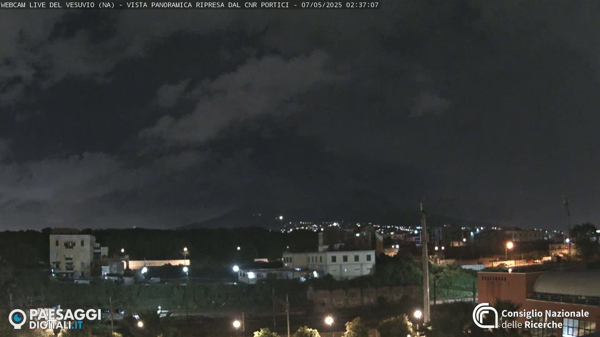A nighttime view of a city with a dark, cloudy sky and a mountain silhouetted in the distance.