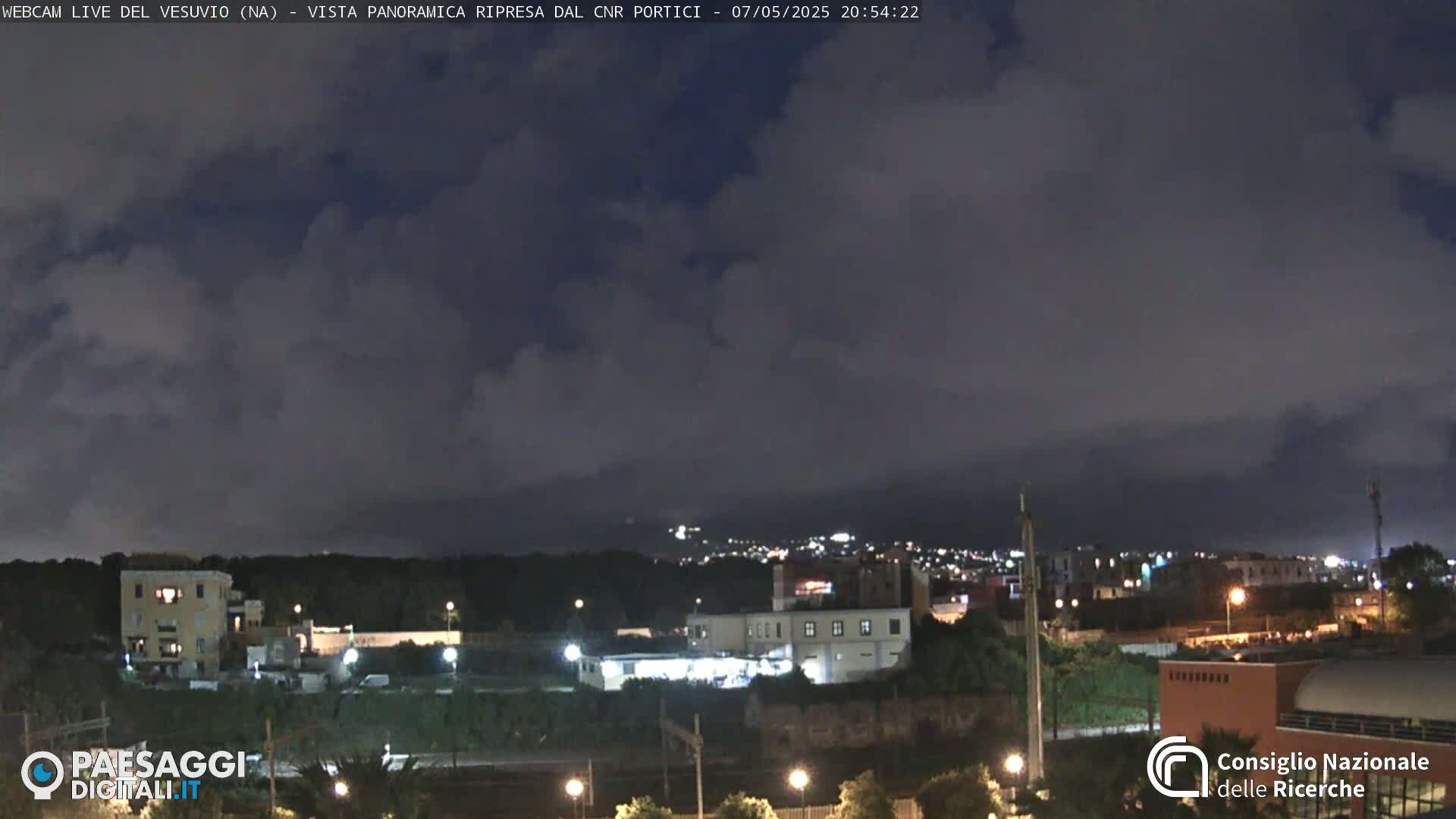 A nighttime view of a city under a mostly cloudy sky shows buildings and lights nestled among dark hills.