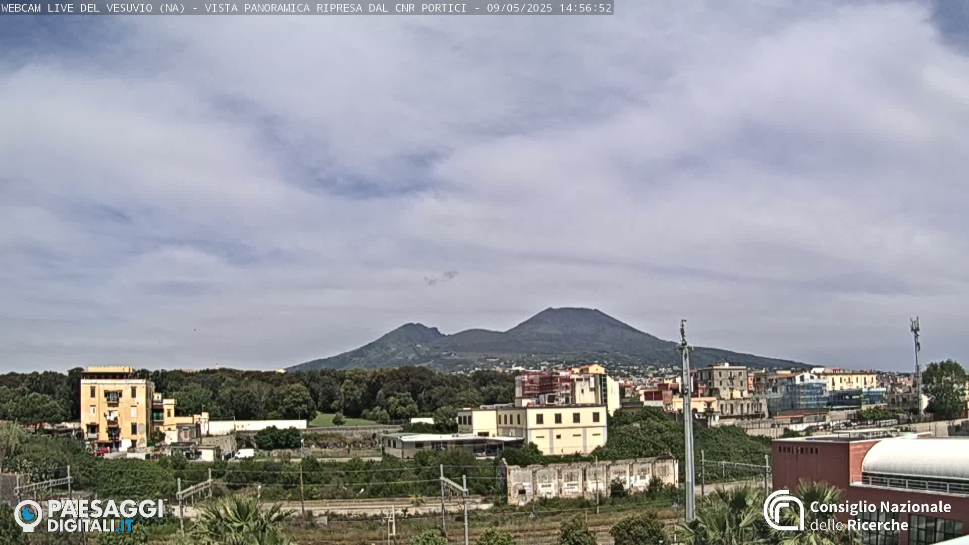 A view of Mount Vesuvius in the distance, under a mostly cloudy sky, with suburban buildings and greenery in the foreground.