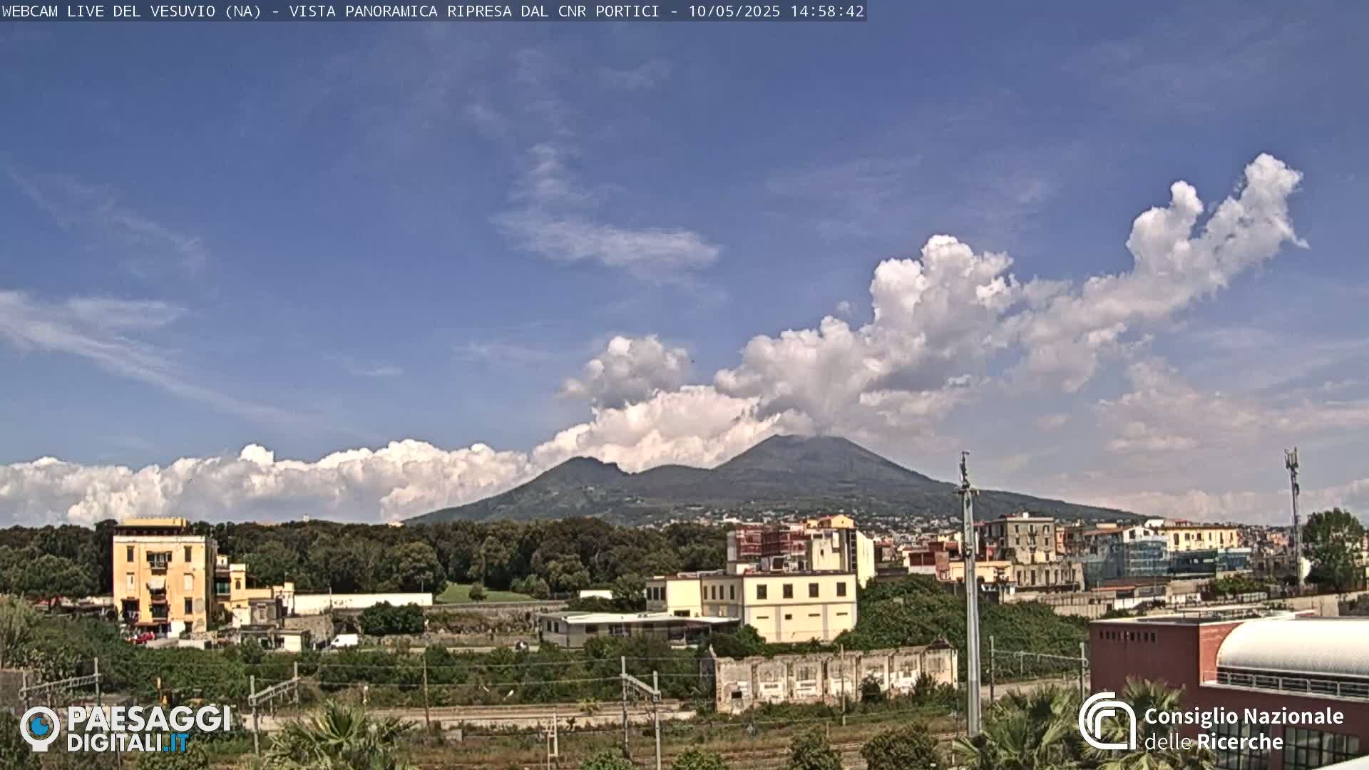 A partly cloudy sky shows Mount Vesuvius in the distance behind a cityscape and green landscape.