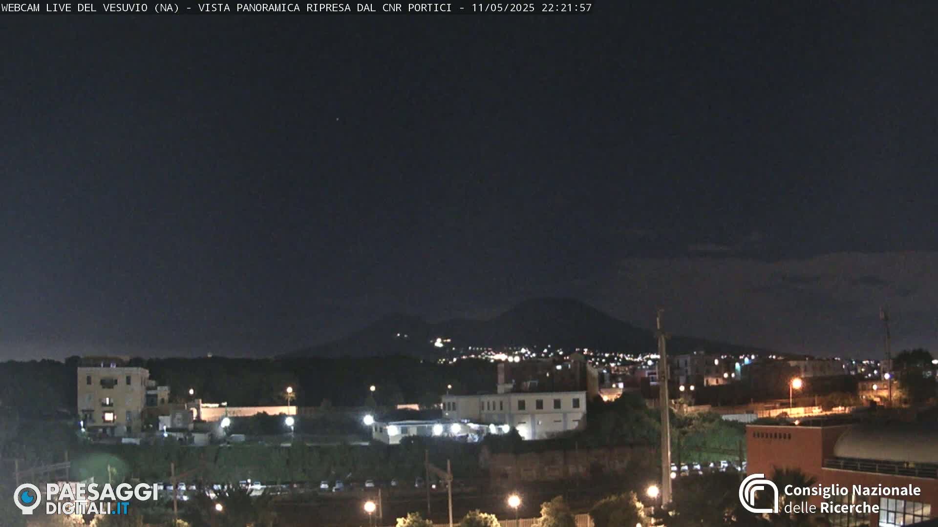 A nighttime view of a city illuminated by lights, with a dark mountain range visible in the distance under a clear, dark sky.